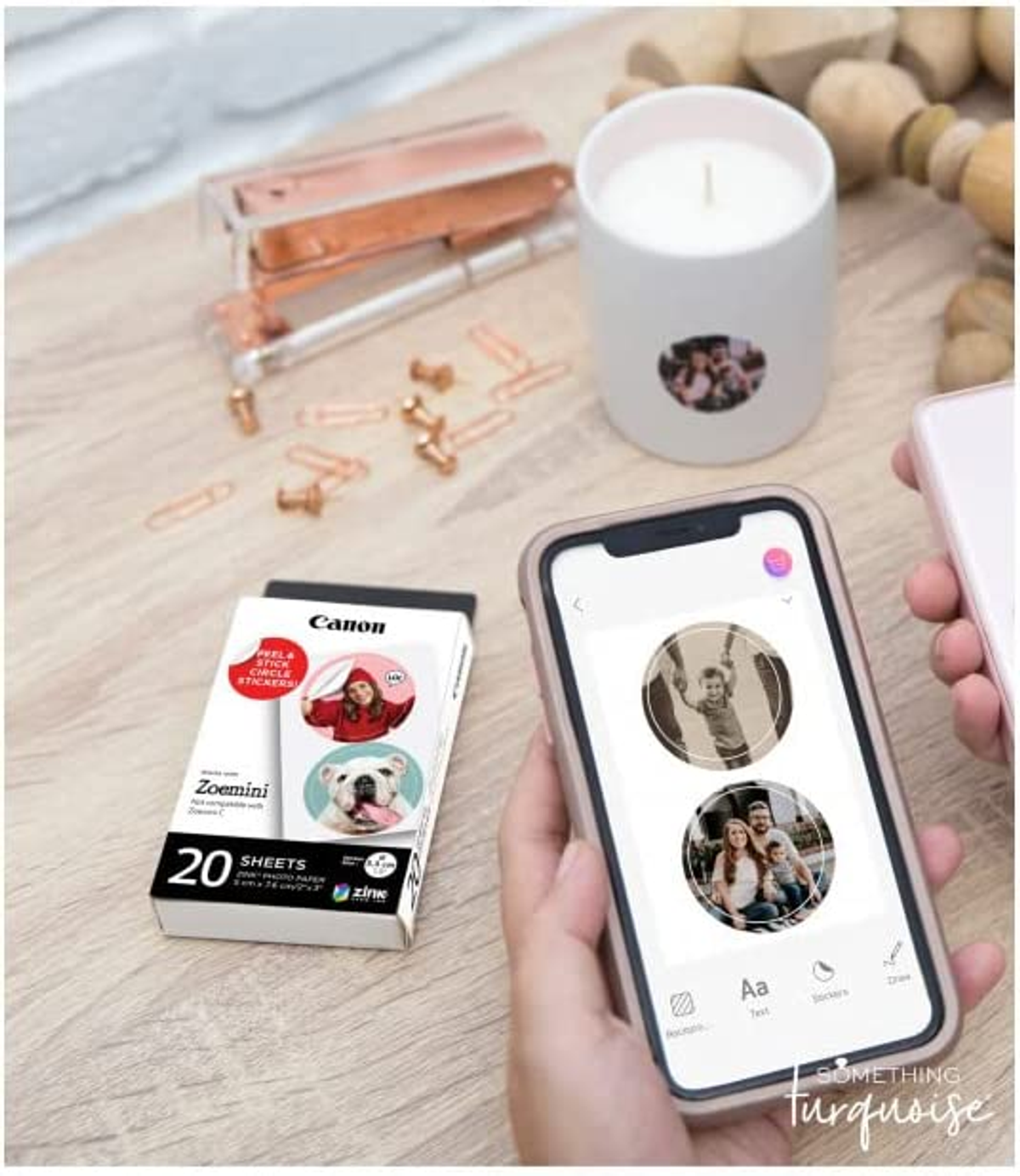 Manos sosteniendo un teléfono con fotos circulares, caja de pegatinas Canon Zoemini y otros objetos.