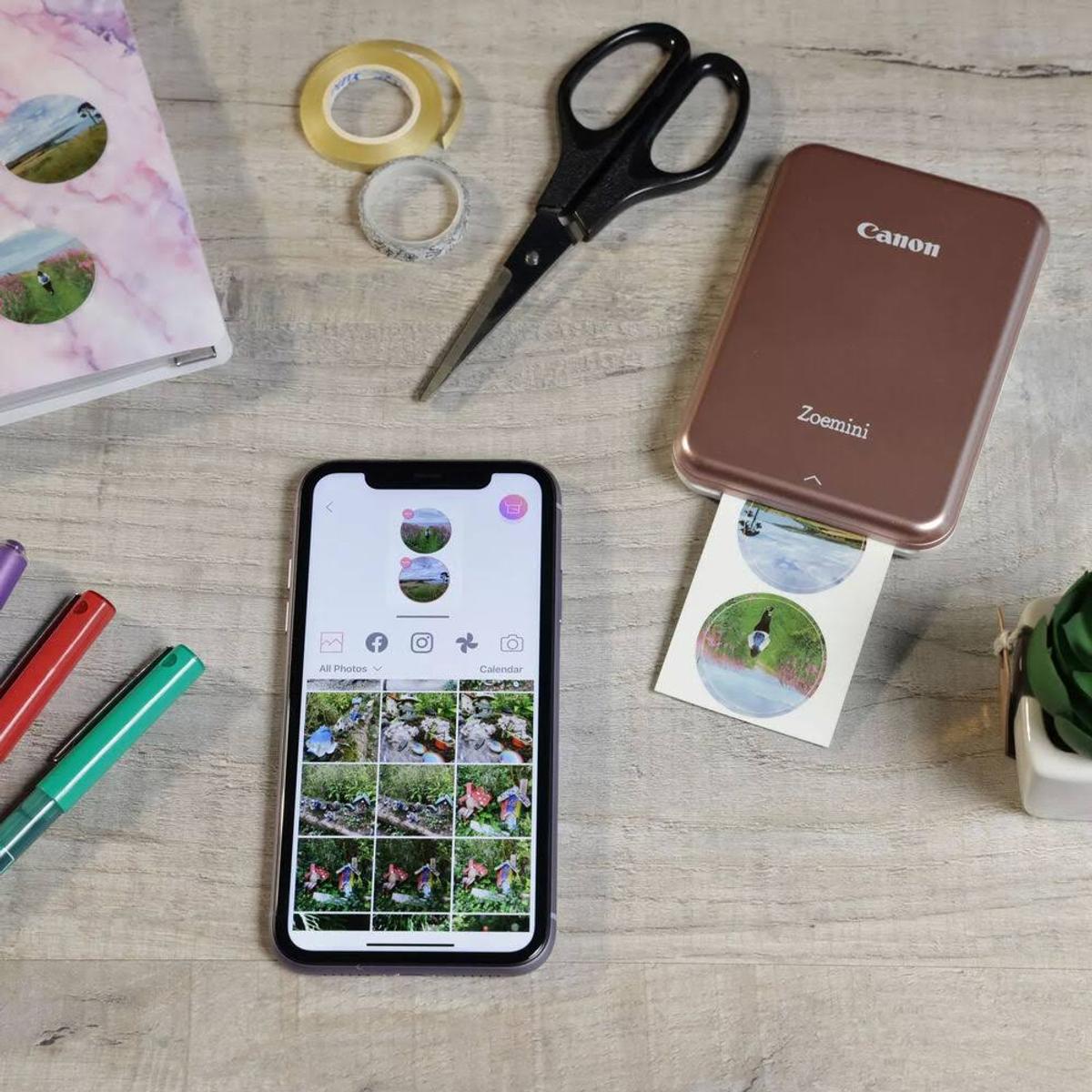 El teléfono muestra fotos, imprime una foto adhesiva con una impresora Canon Zoemini. Tijeras y cinta en una mesa.