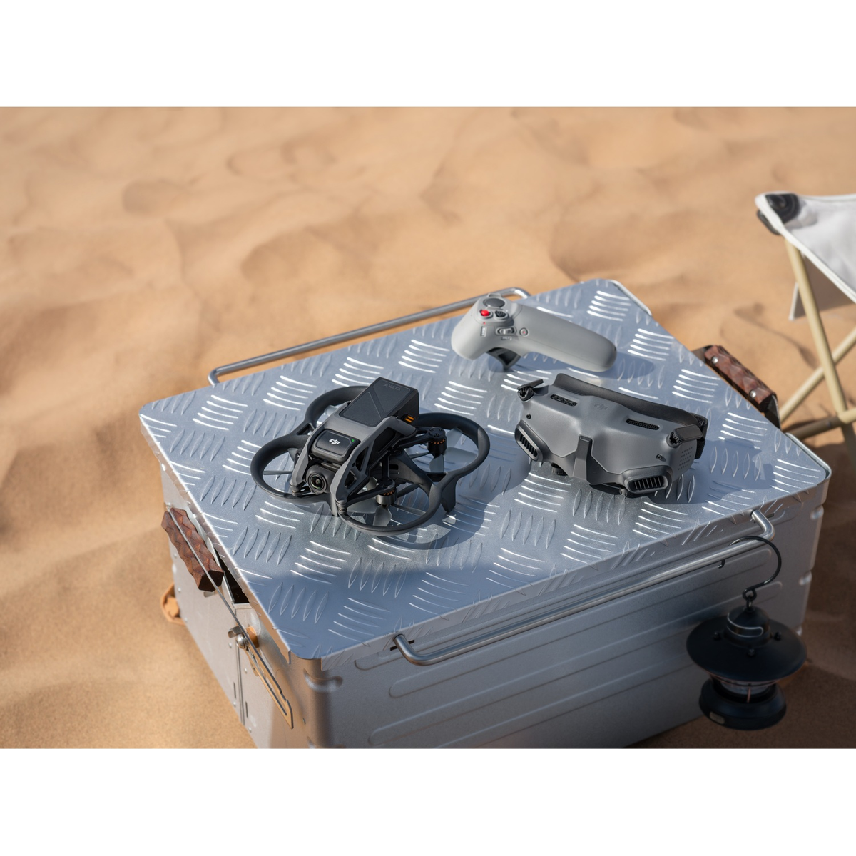 Dron y controlador sobre una caja plateada en un desierto con silla de camping y linterna.