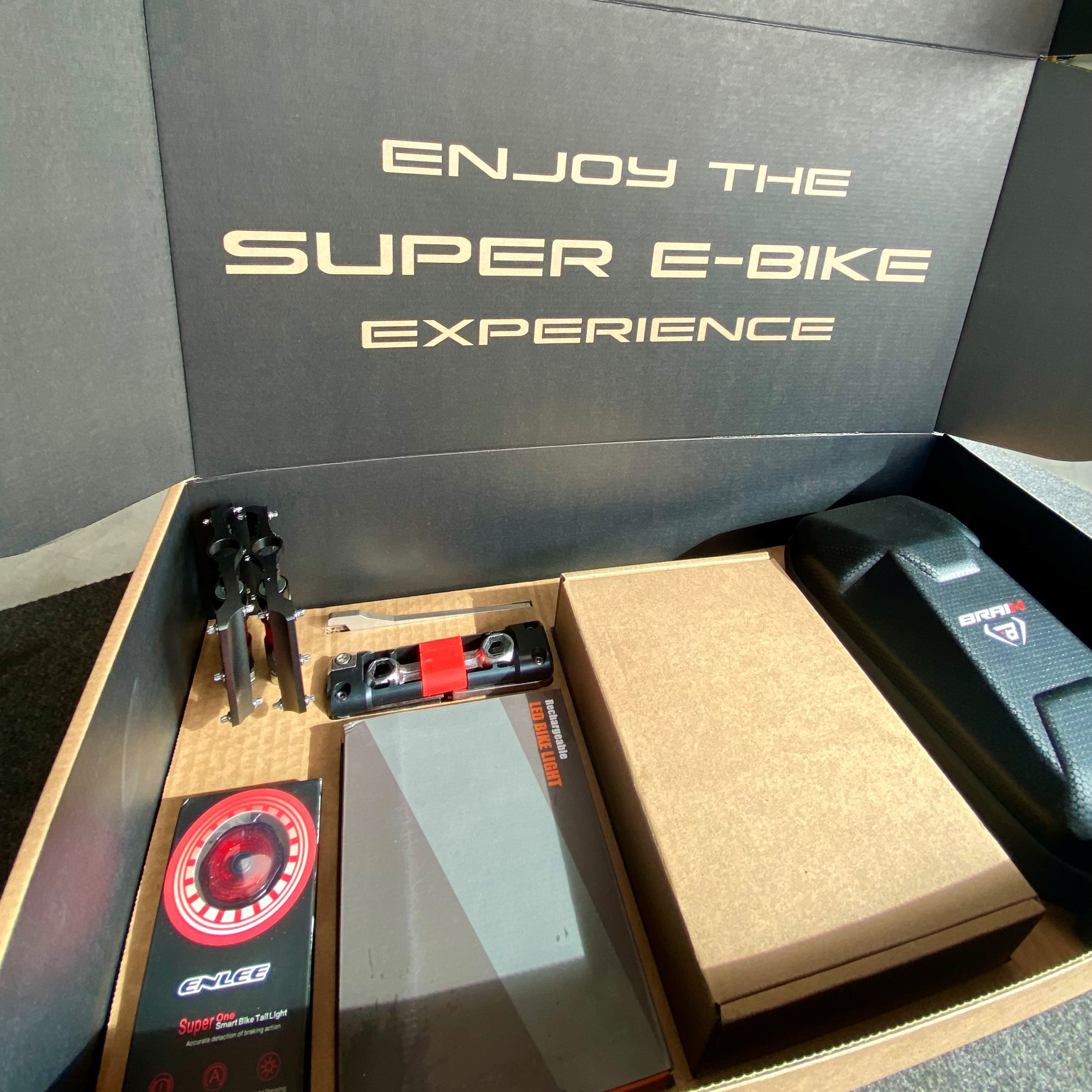 Caja de cartón abierta con accesorios para e-bike. Texto: 'ENJOY THE SUPER E-BIKE EXPERIENCE'.