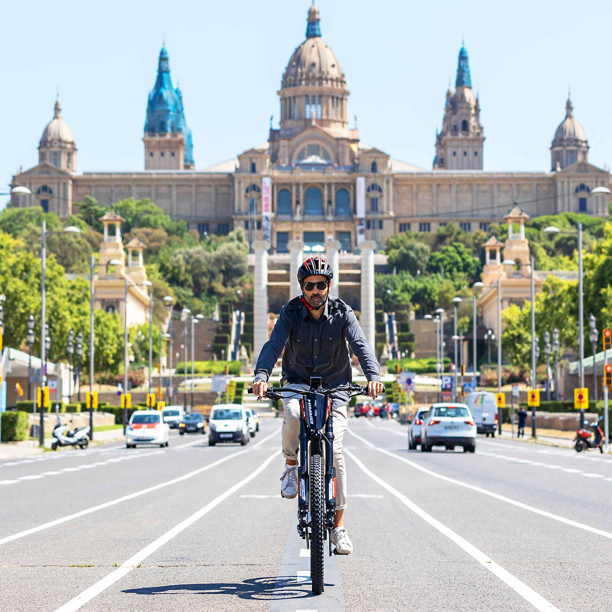 Un hombre en bicicleta pedaleando hacia la cámara frente a un edificio y una carretera.