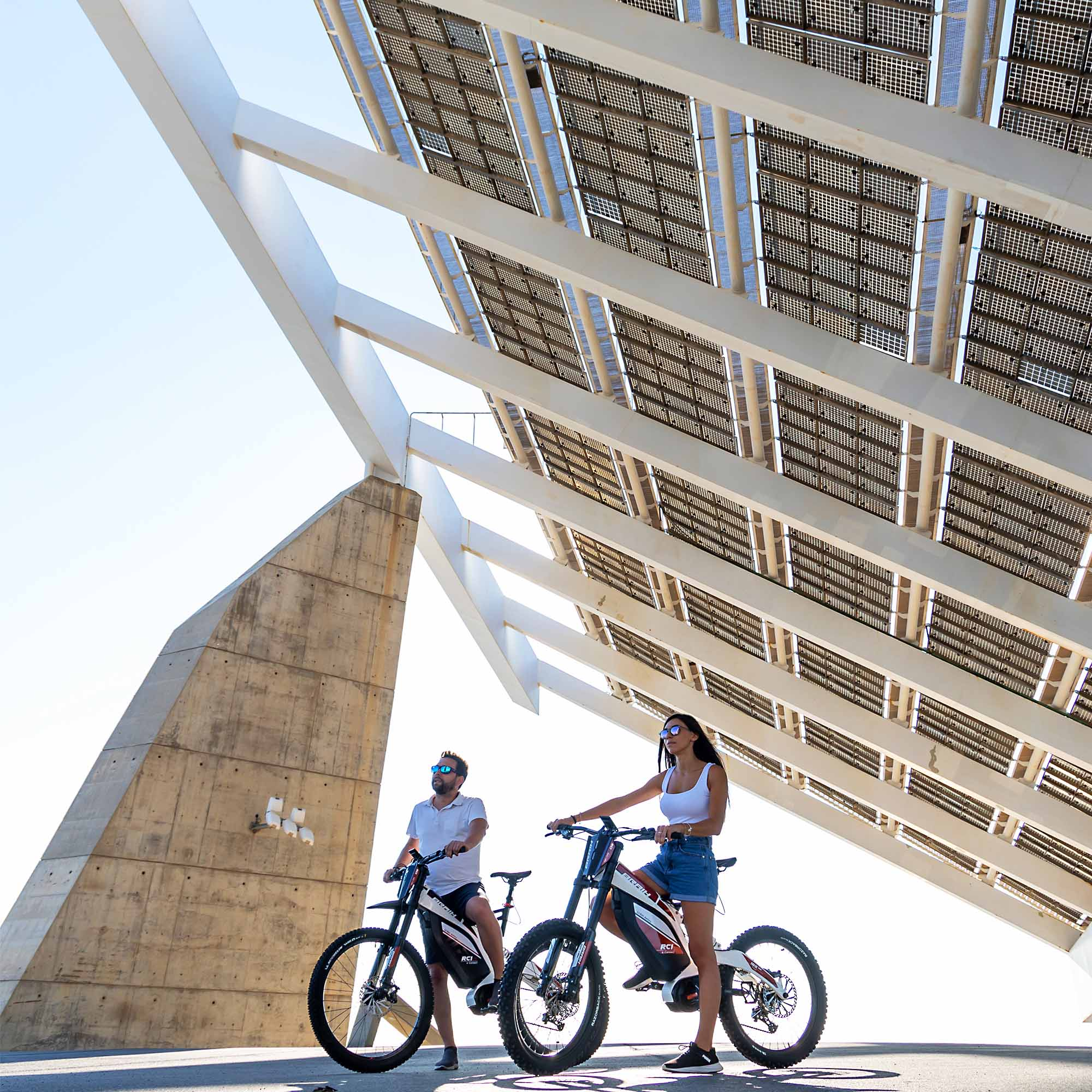 Dos personas en bicicletas eléctricas frente a una estructura de paneles solares, día soleado.