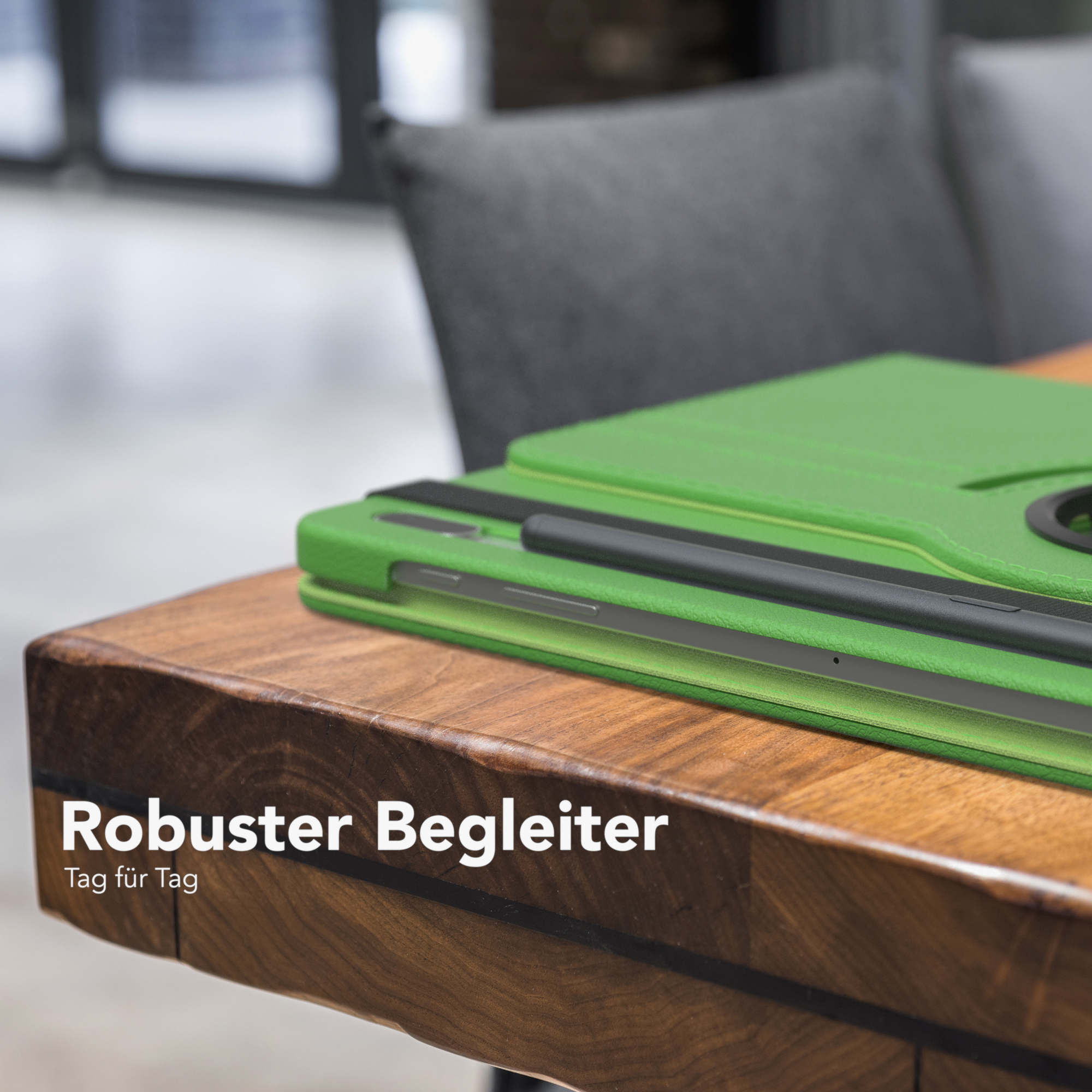Grüne Tablethülle auf einem Holztisch. Text im Bild lautet 'Robuster Begleiter'. Grauer Stuhl im Hintergrund.