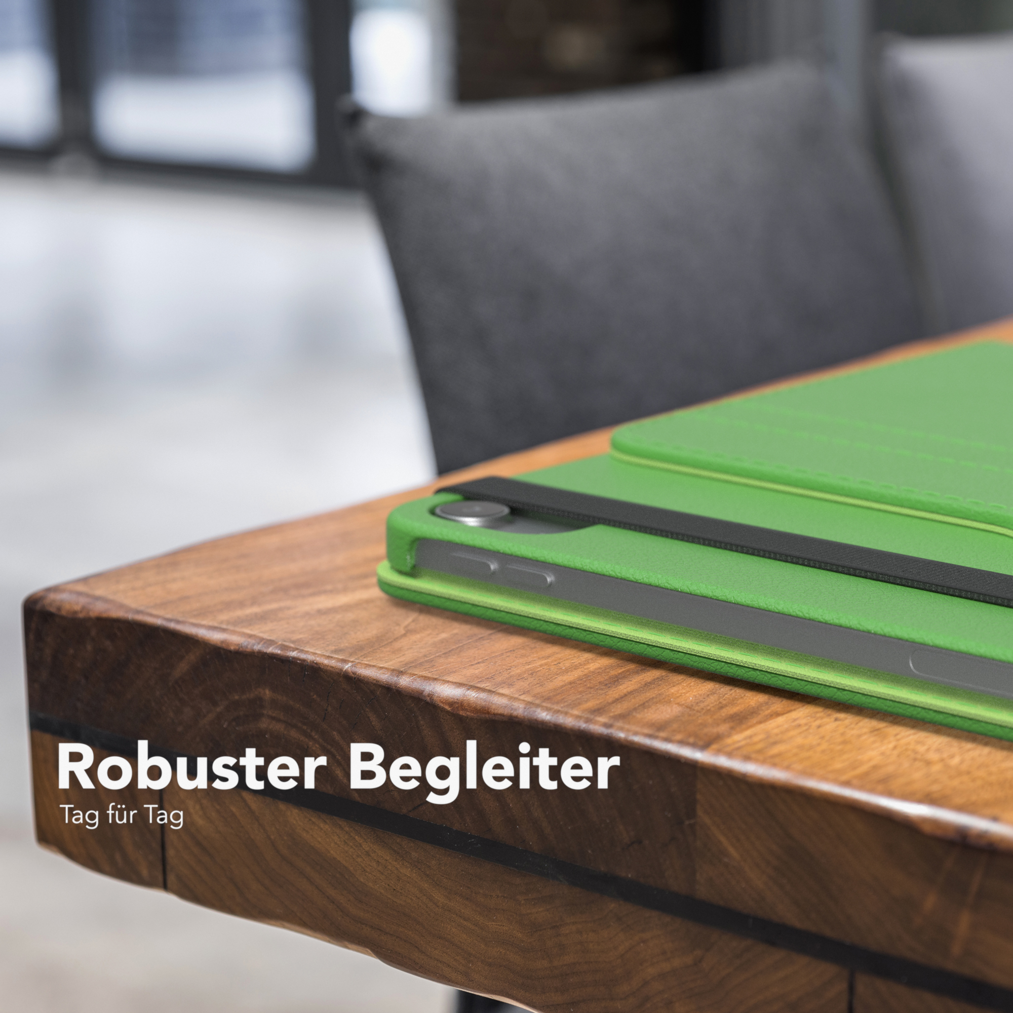 Grüne Tablettasche auf Holztisch. Text auf Deutsch. Hintergrund: grauer Stuhl.