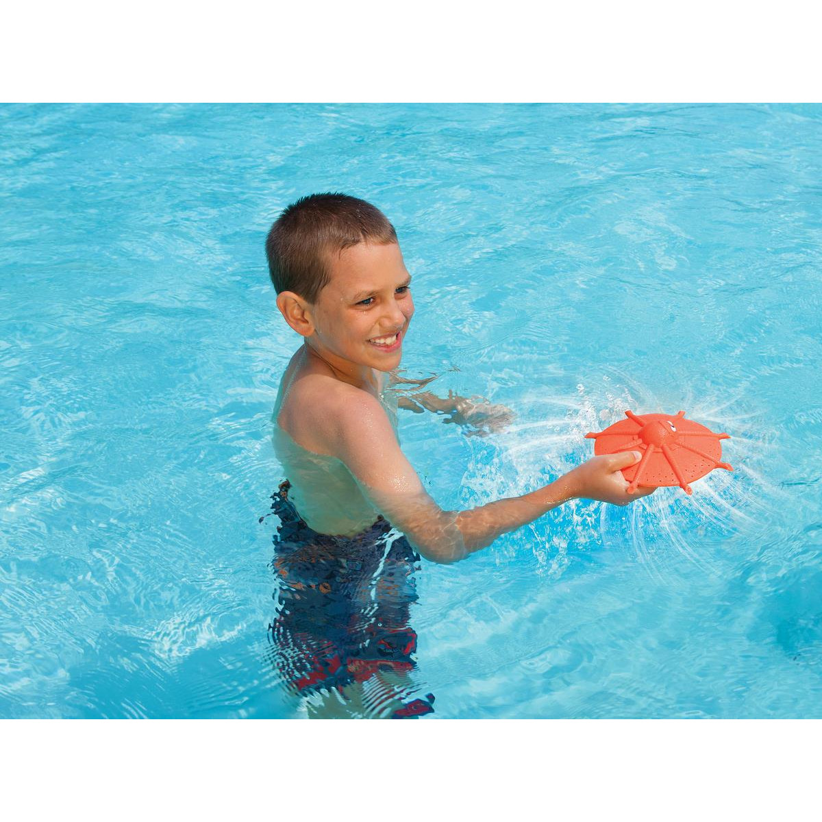 Junge in einem Pool, der ein orangefarbenes Wasserspielzeug hält. Das Spielzeug spritzt Wasser. Der Junge lächelt.