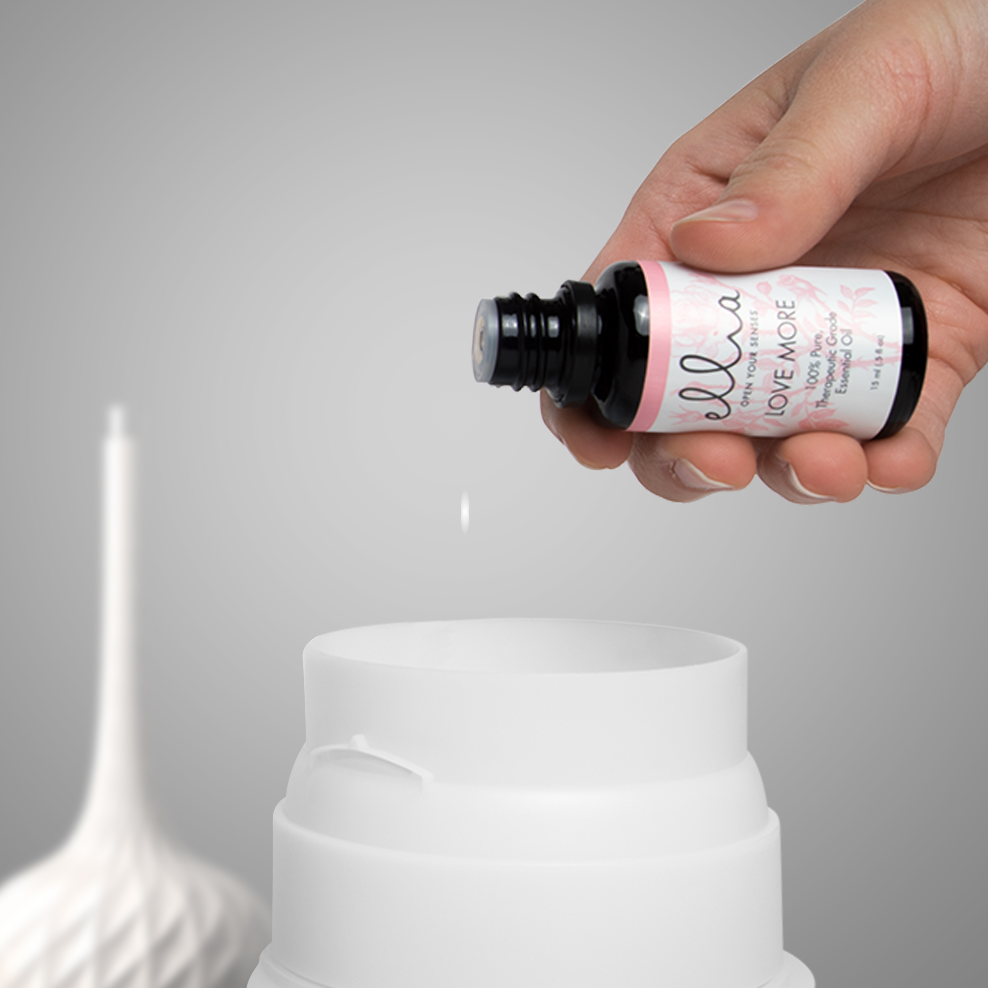 Mano che versa olio essenziale in un diffusore. Diffusore bianco e bottiglia con etichetta LOVE MORE.