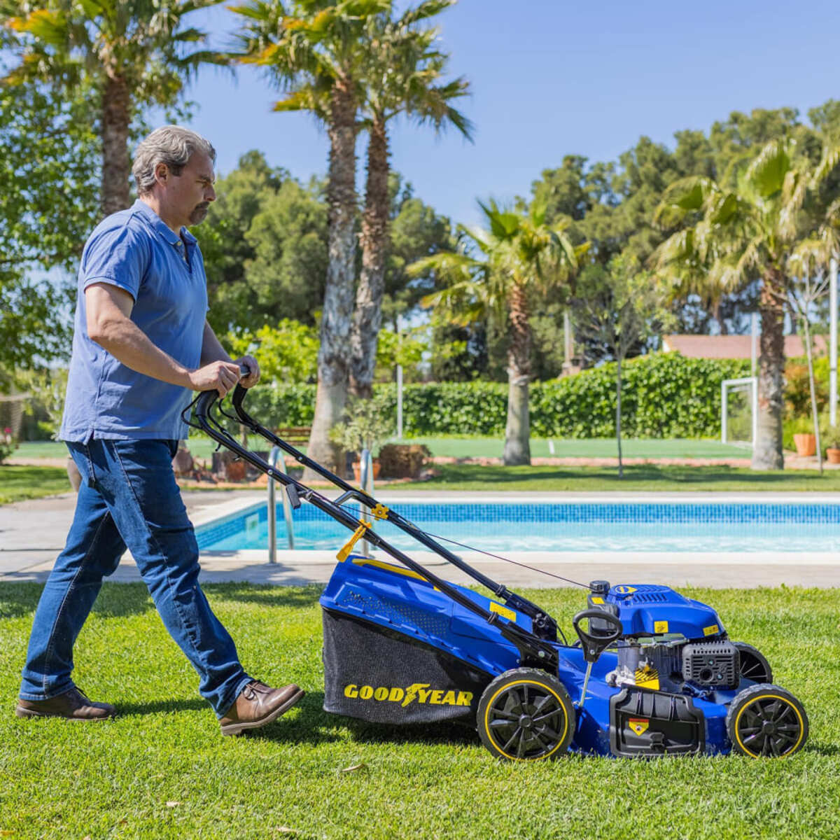 Un hombre empuja un cortacésped azul y negro sobre hierba verde cerca de una piscina y palmeras.