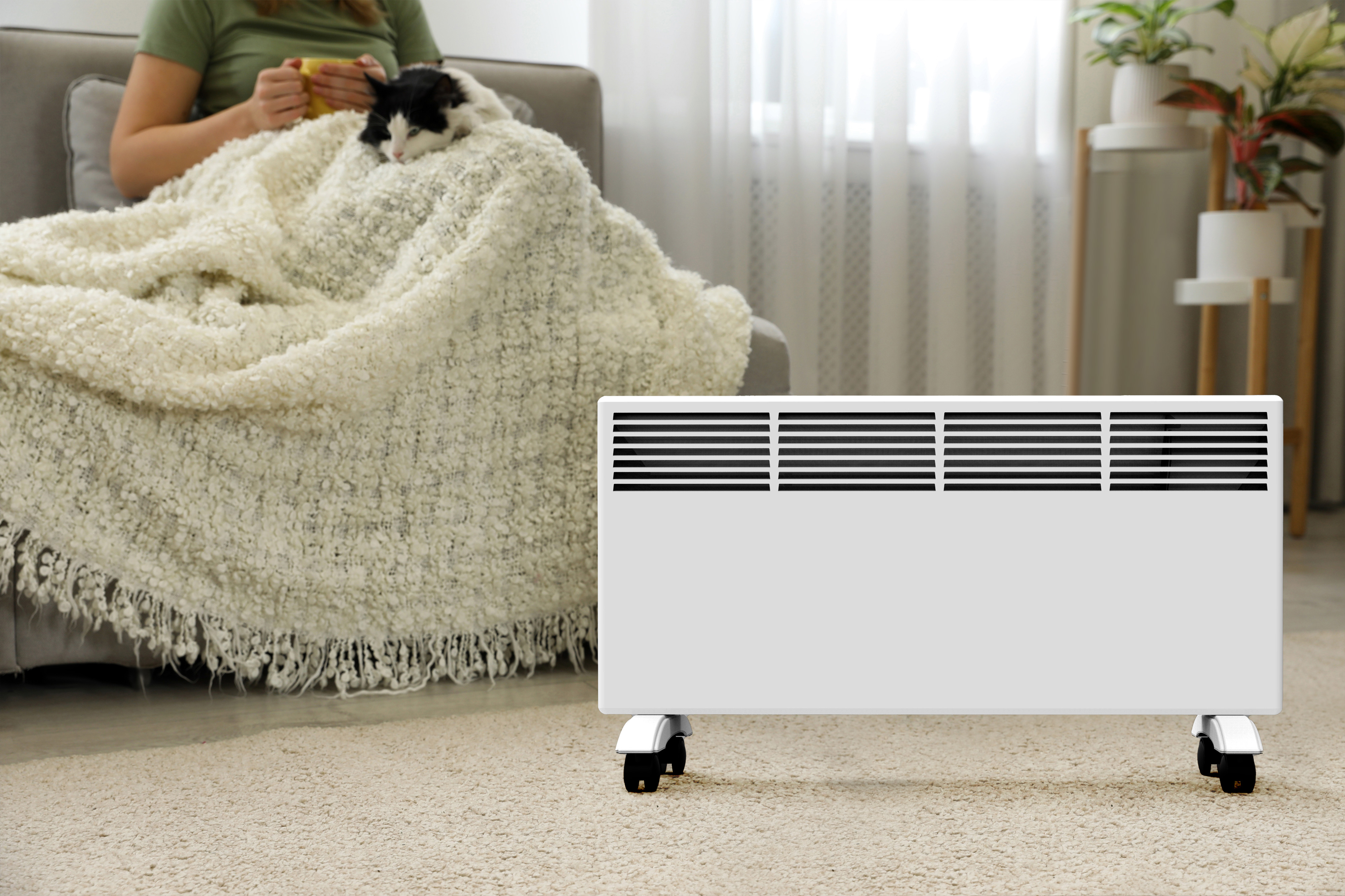 Weißer Elektroheizer auf Rollen. Im Hintergrund sitzt eine Frau mit einer Katze auf einem Sofa.