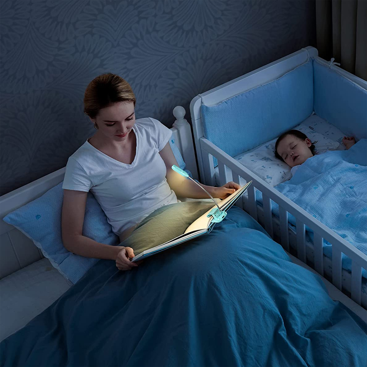 Eine Frau liest mit einer Leselampe, ein Baby schläft in einem Kinderbett in der Nähe.