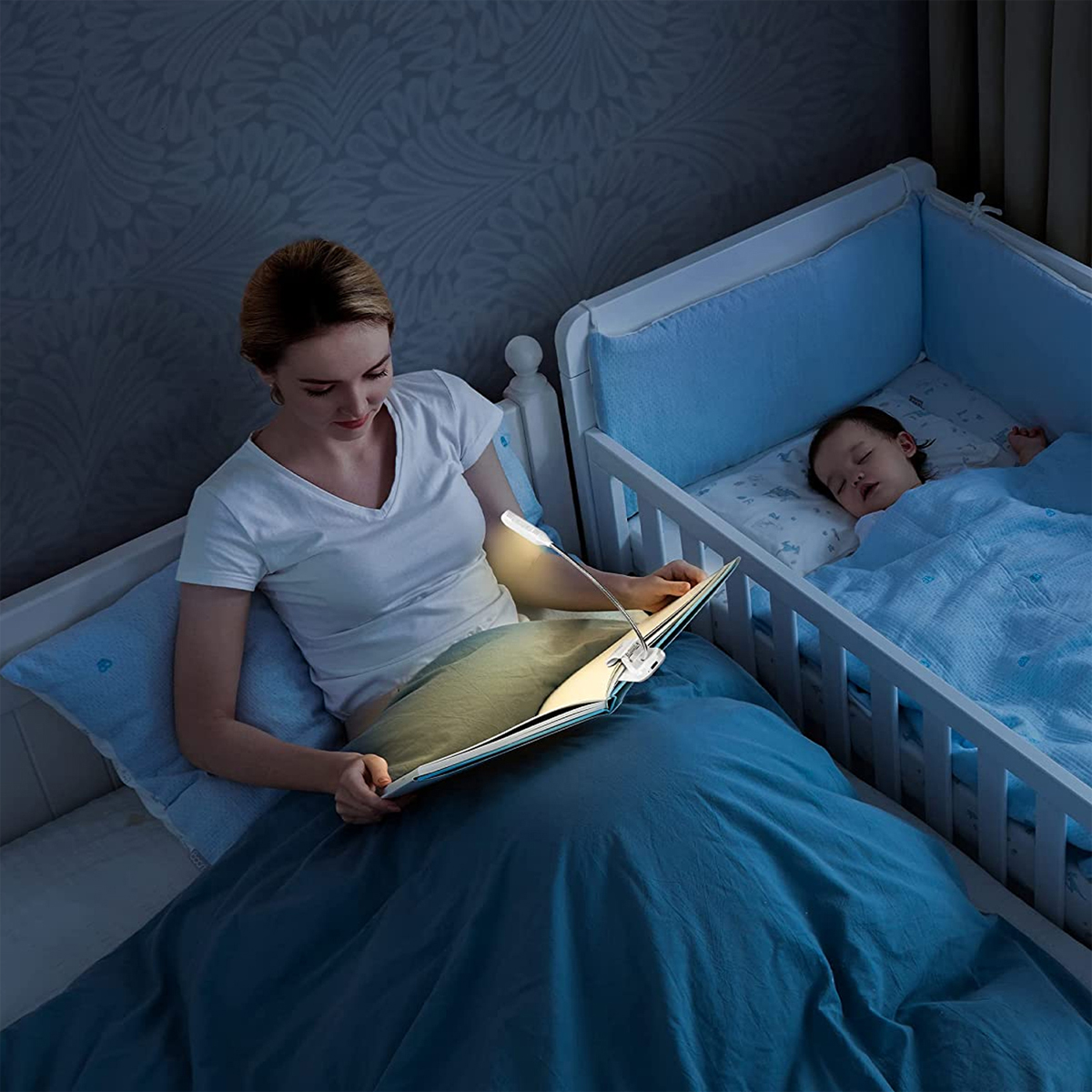 Frau liest im Bett mit Leselampe, Baby schläft in der Nähe im Kinderbett.