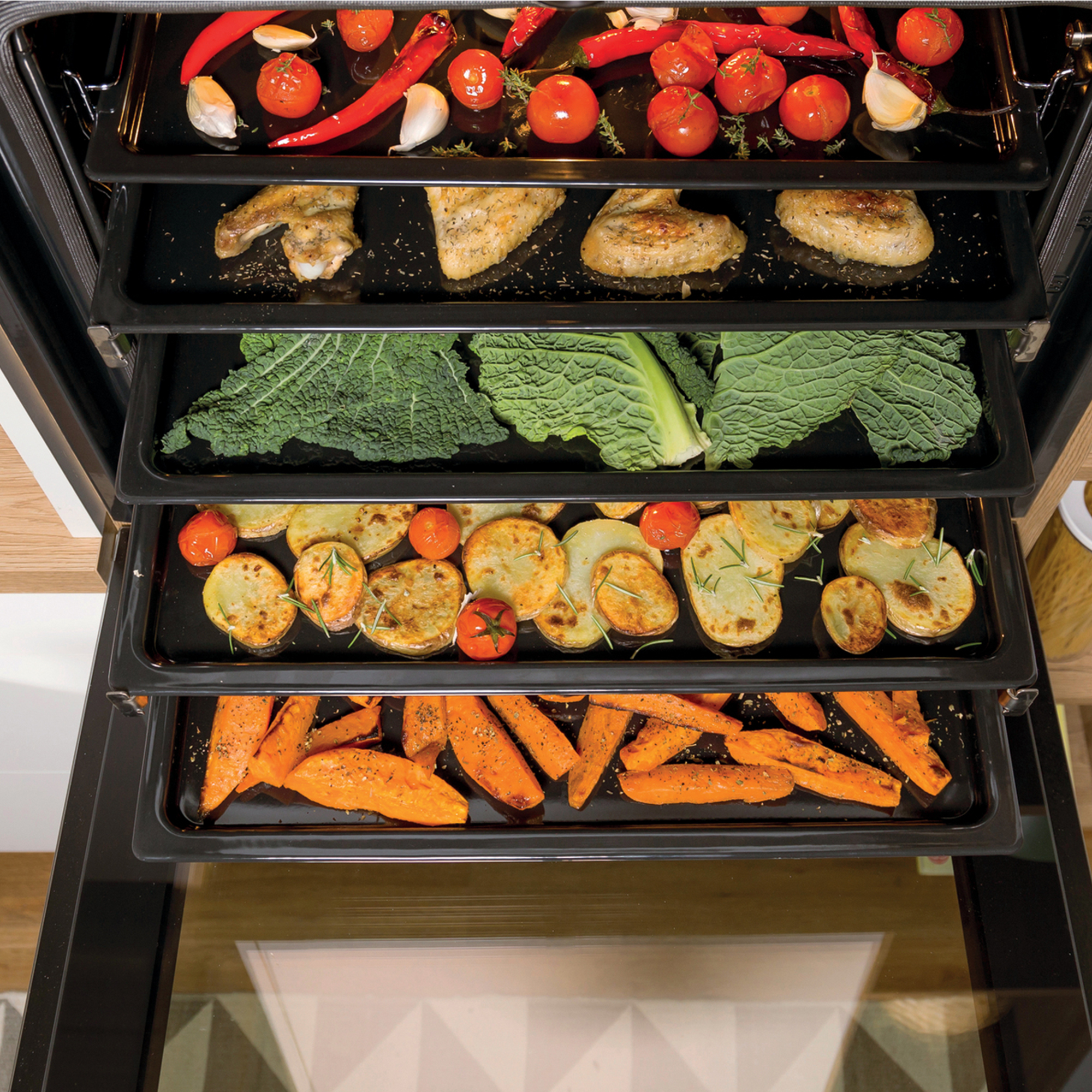 Piekarnik z tacami z jedzeniem. Górna taca ma skrzydełka i warzywa. Dolna taca ma bataty.