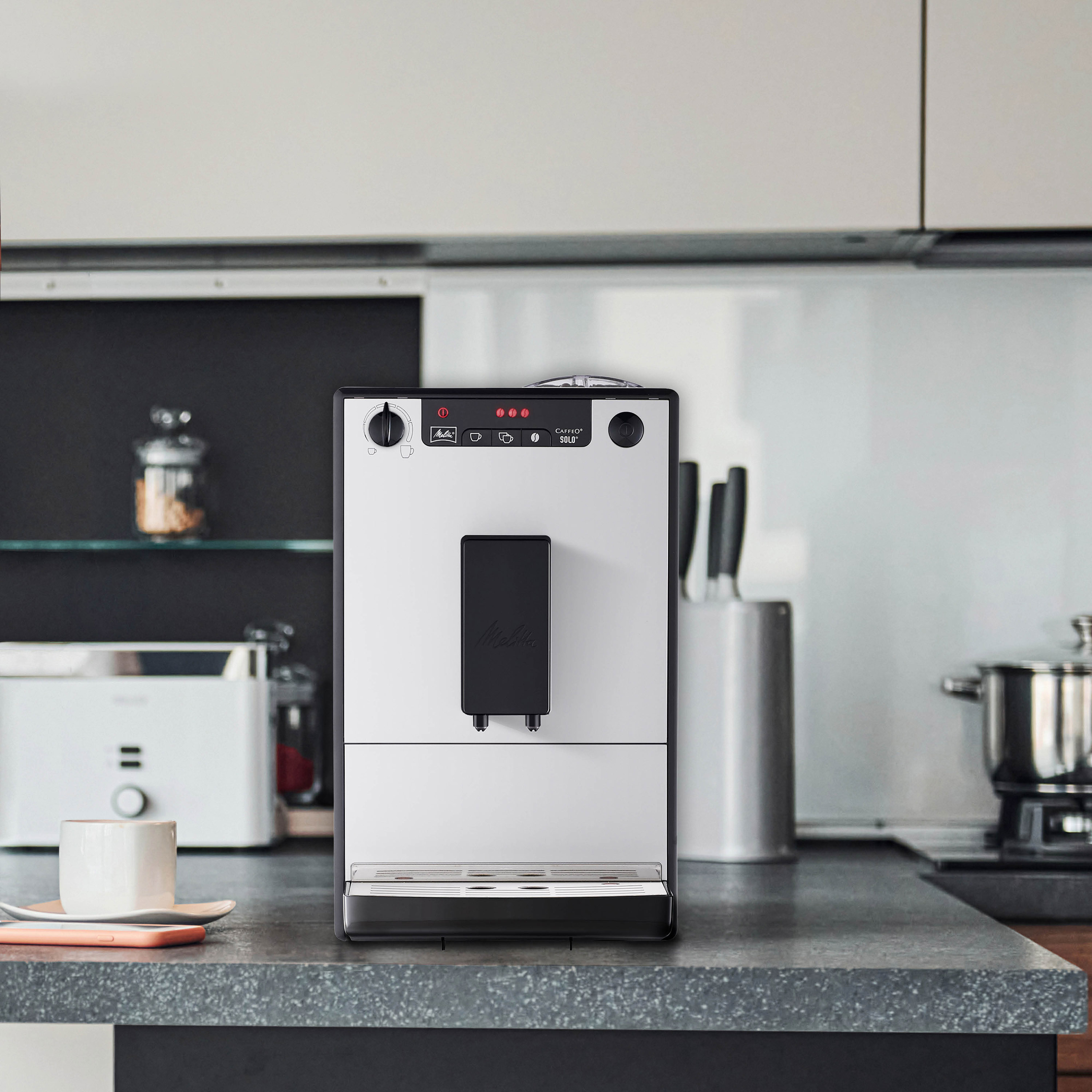 Biały i srebrny ekspres do kawy. Czarny przód, z filiżanką kawy i tłem kuchni.