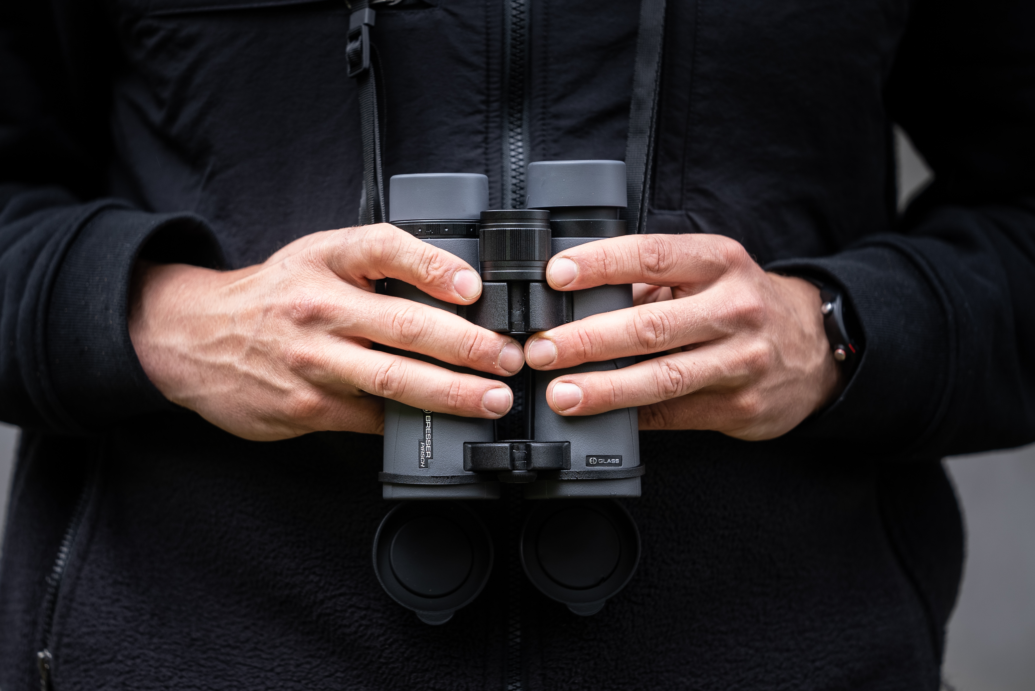 Person hält Fernglas. Das Fernglas ist grau und schwarz. Die Person trägt eine dunkle Jacke.