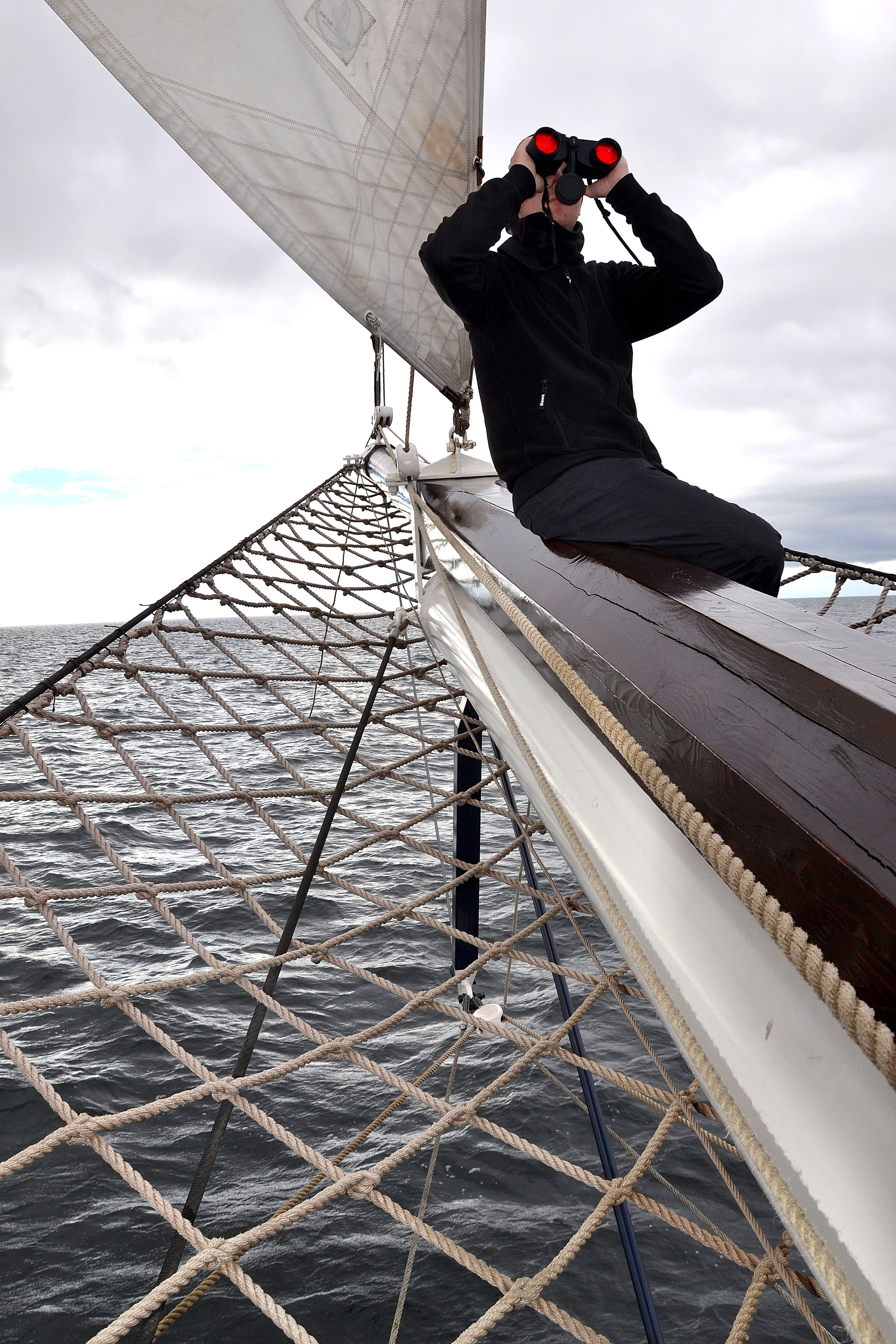 Hombre en un velero mirando a través de binoculares. El cielo está nublado y el agua picada.