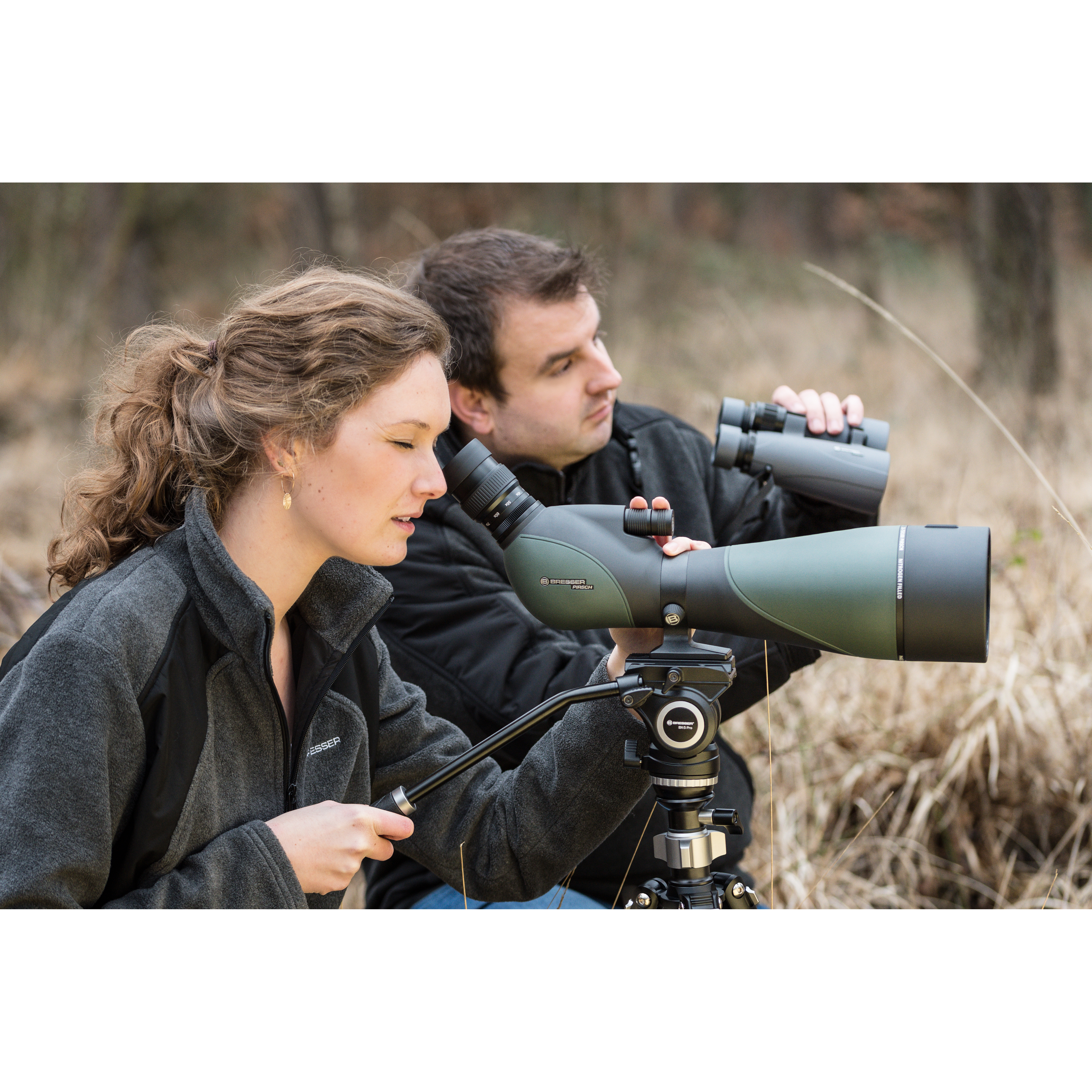 Un hombre y una mujer usando un telescopio terrestre con binoculares, al aire libre. Están en un campo, enfocados en algo.