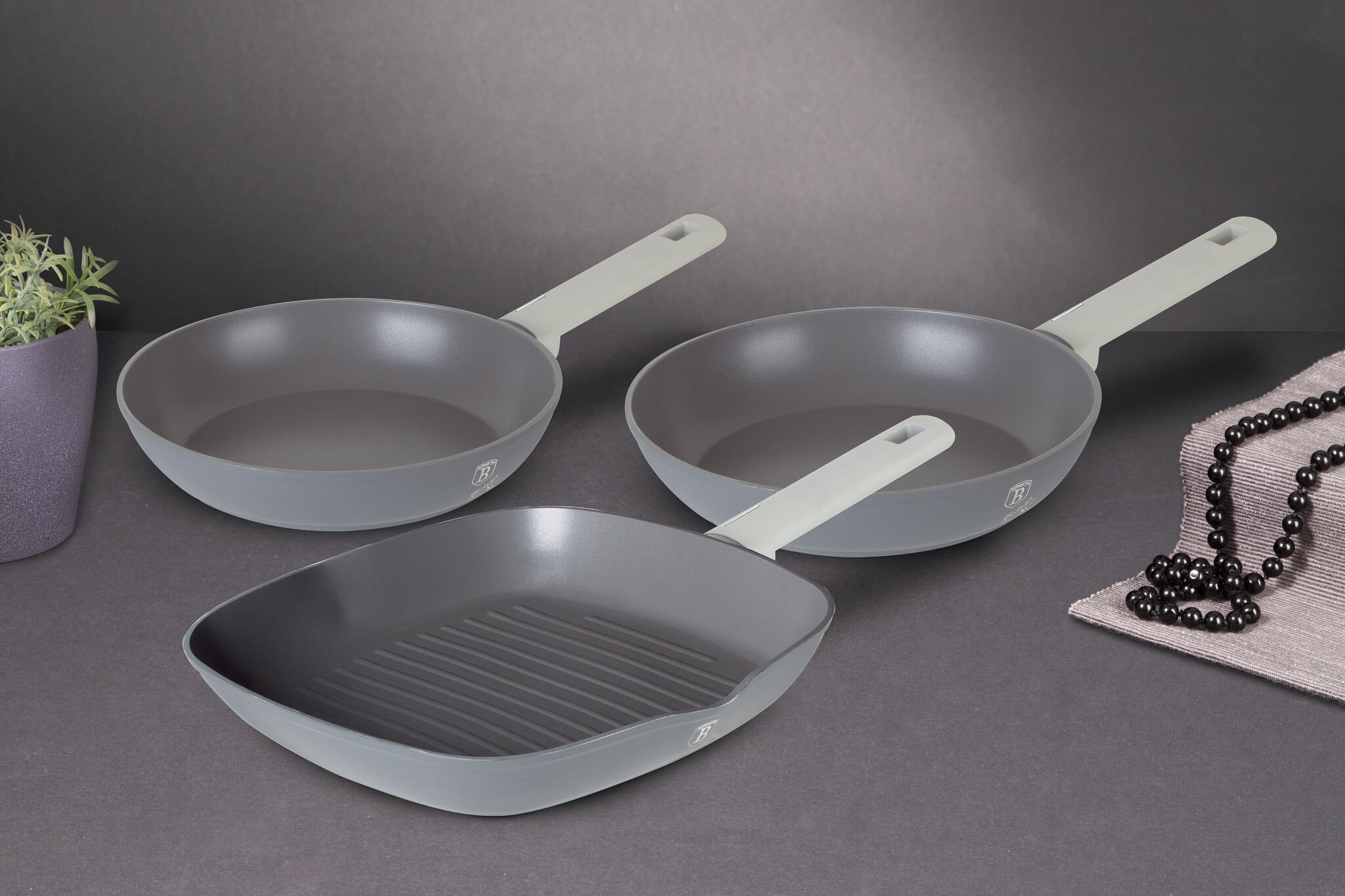 Drei graue Bratpfannen mit weißen Griffen. Eine ist quadratisch mit Grilllinien. Dunkelgrauer Hintergrund.