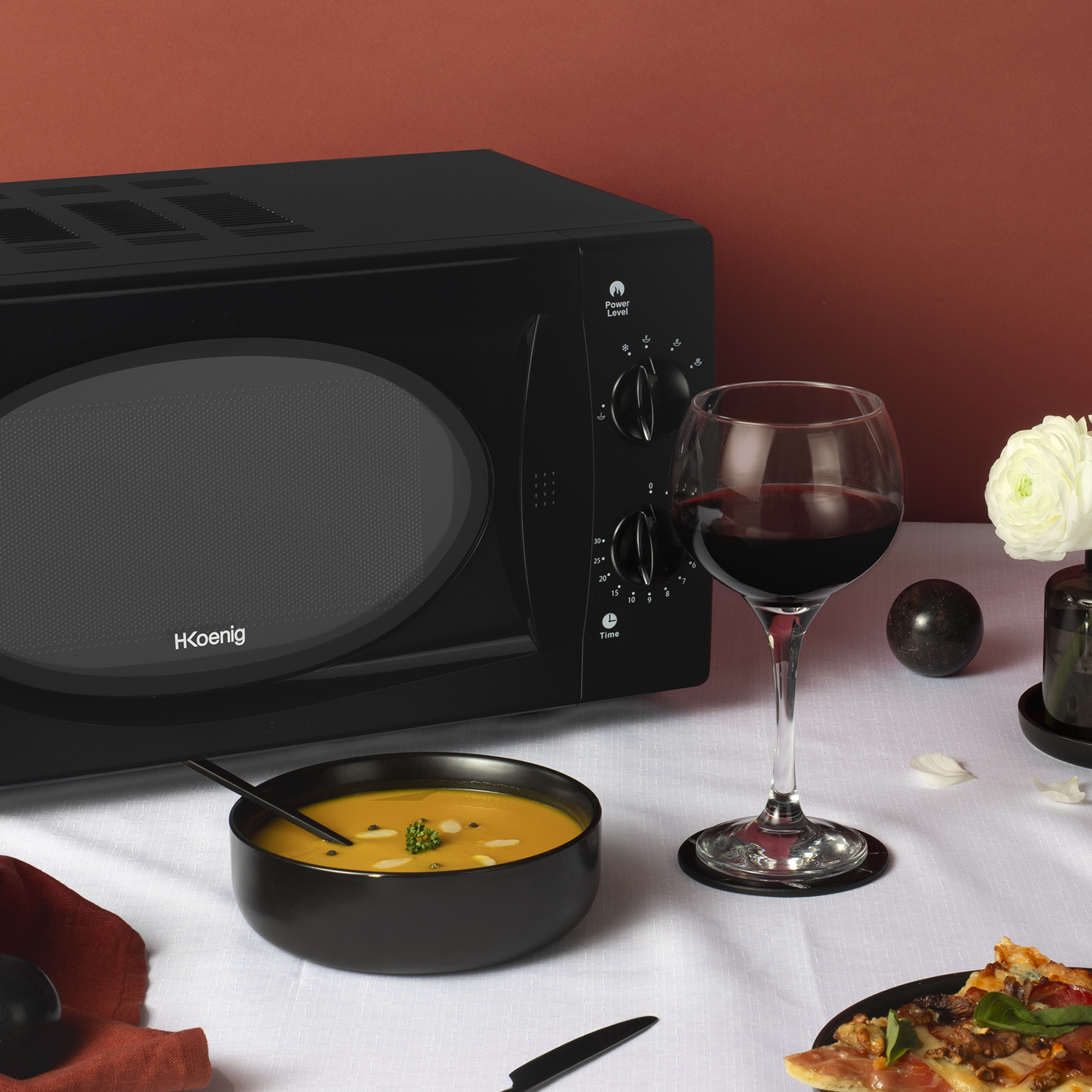 Microonde nero, ciotola di zuppa, bicchiere di vino con vino rosso e una pizza su una tovaglia bianca.