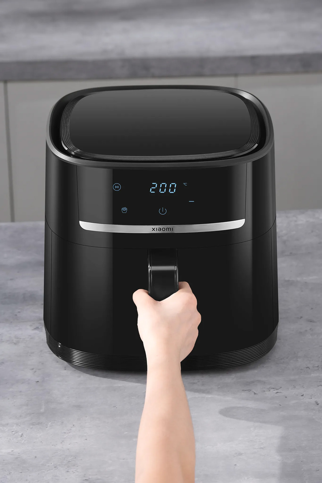 Czarna frytkownica powietrzna z wyświetlaczem, dłoń na uchwycie. Szare tło.