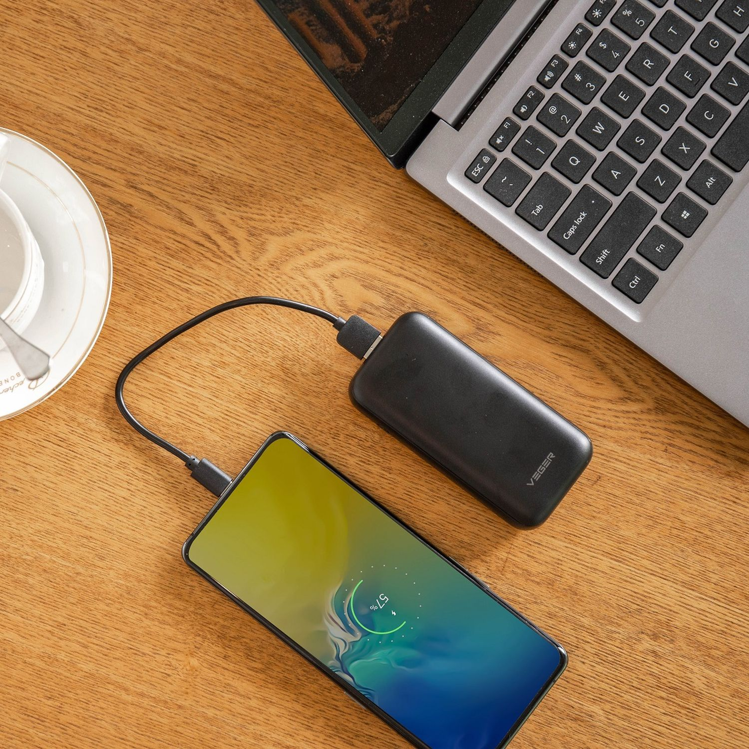 Telefono in carica con power bank su tavolo in legno, vicino a laptop e tazza di caffè.