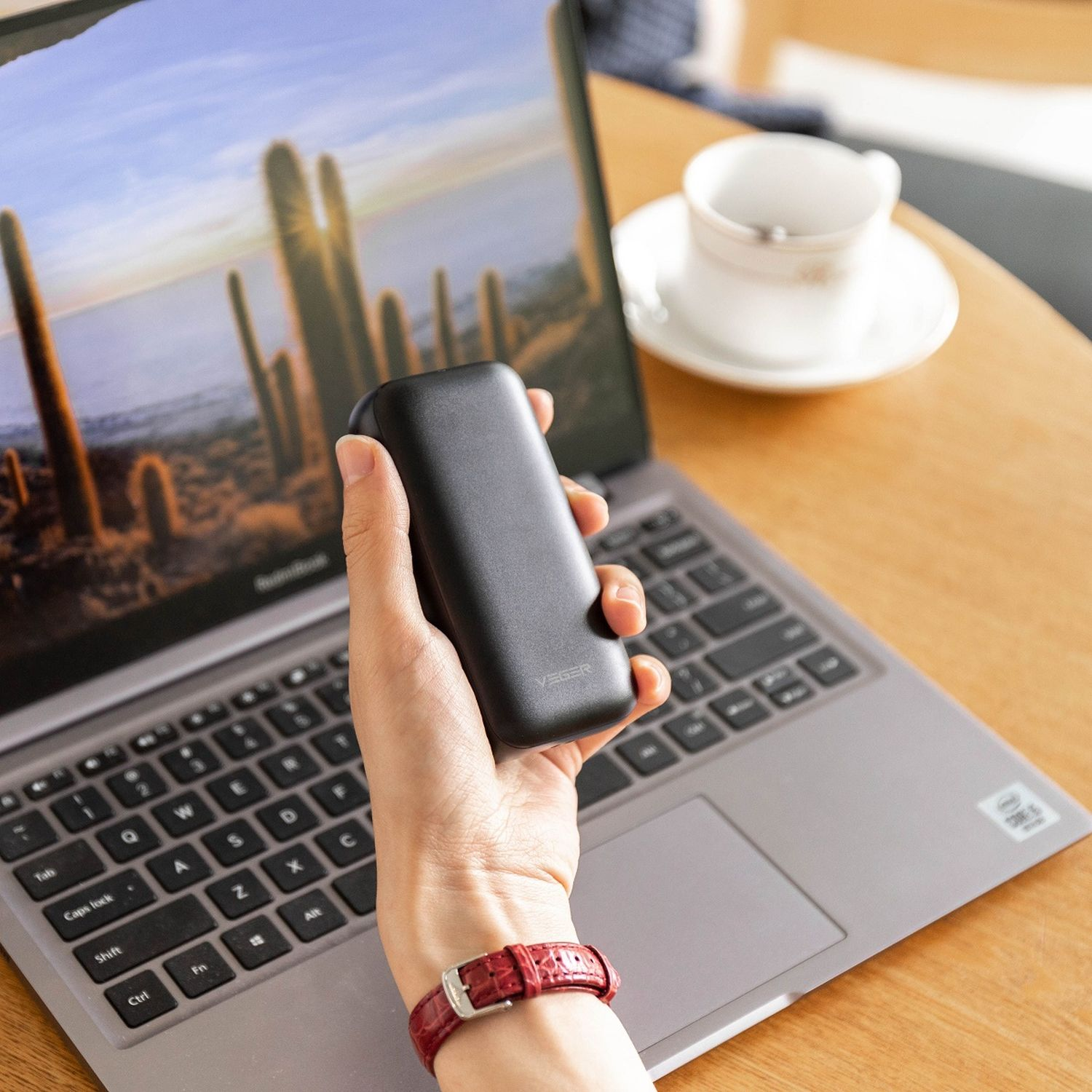 Una persona tiene in mano un dispositivo nero, vicino a un laptop. Una tazza di caffè è su un tavolo. È visibile un orologio da polso.
