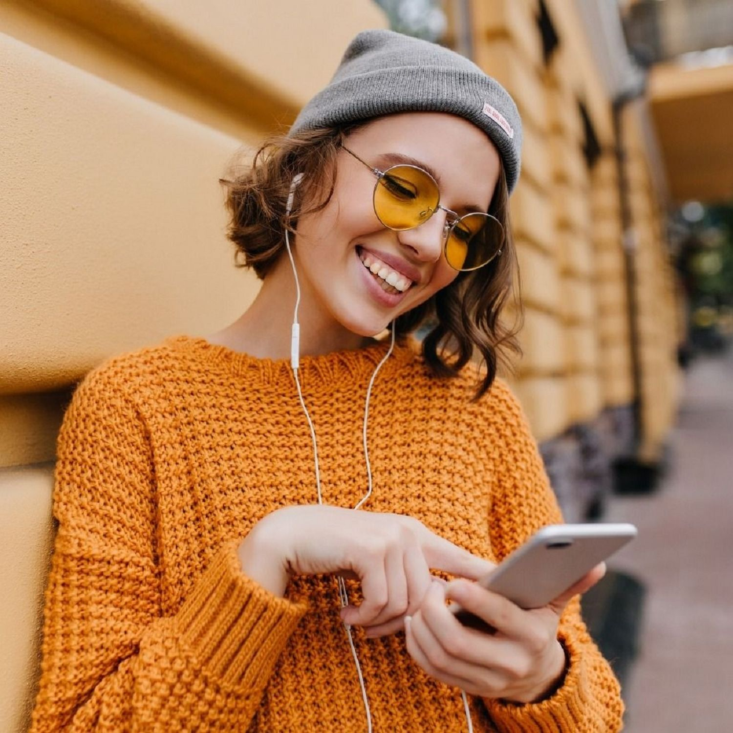 Lächelnde Frau mit Mütze und orangefarbenem Pullover, die ein Telefon benutzt, gelbe Brille tragend.