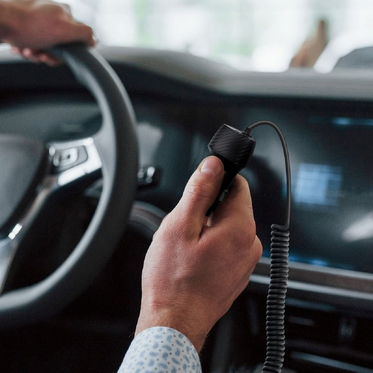 Die Hand einer Person hält ein Autoladegerät in einem Auto. Das Ladegerät ist schwarz und mit einem Kabel verbunden.