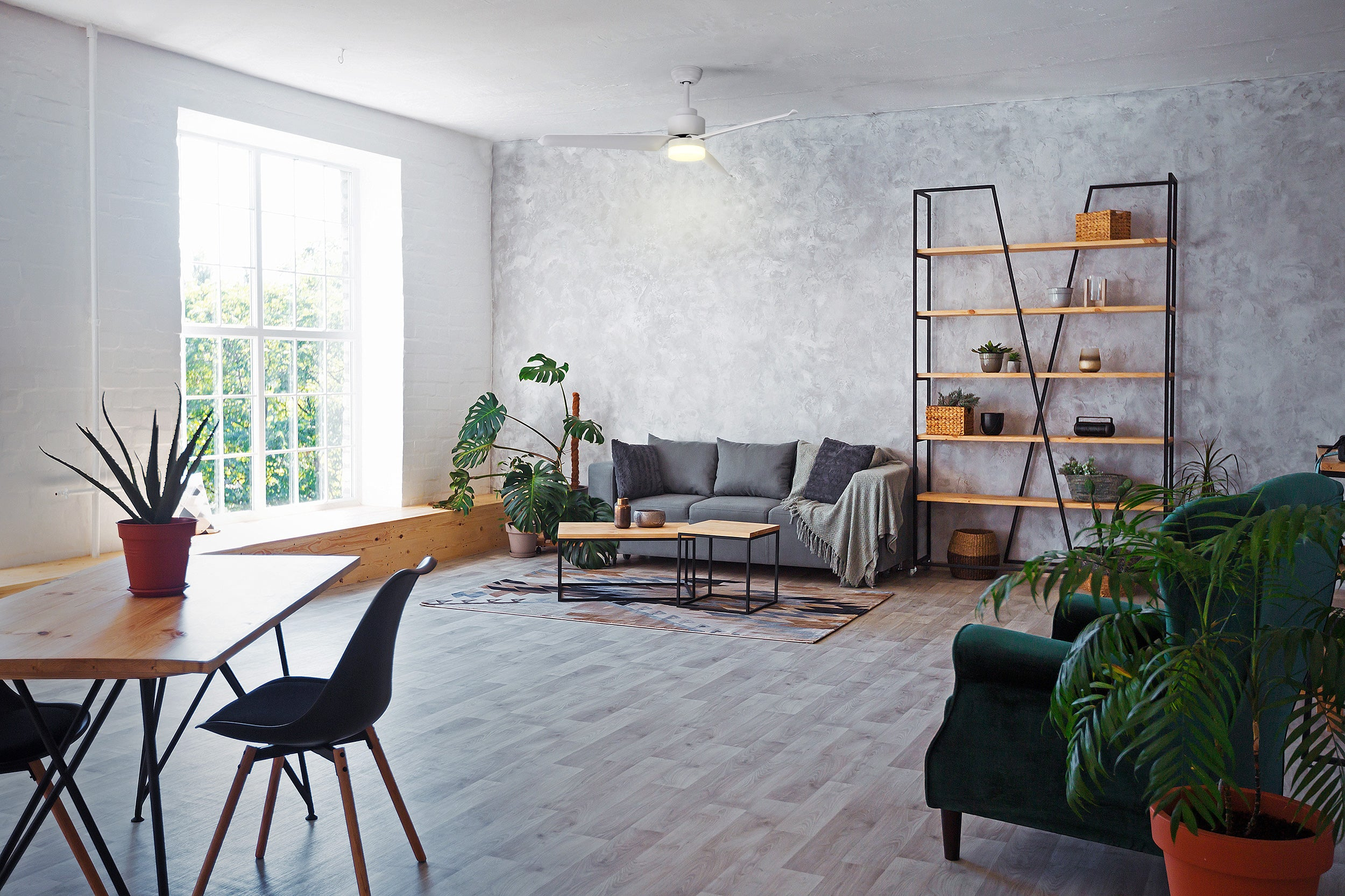 Salon z szarą sofą, drewnianym stołem, roślinami i dużym oknem. Jasna drewniana podłoga i białe ściany.