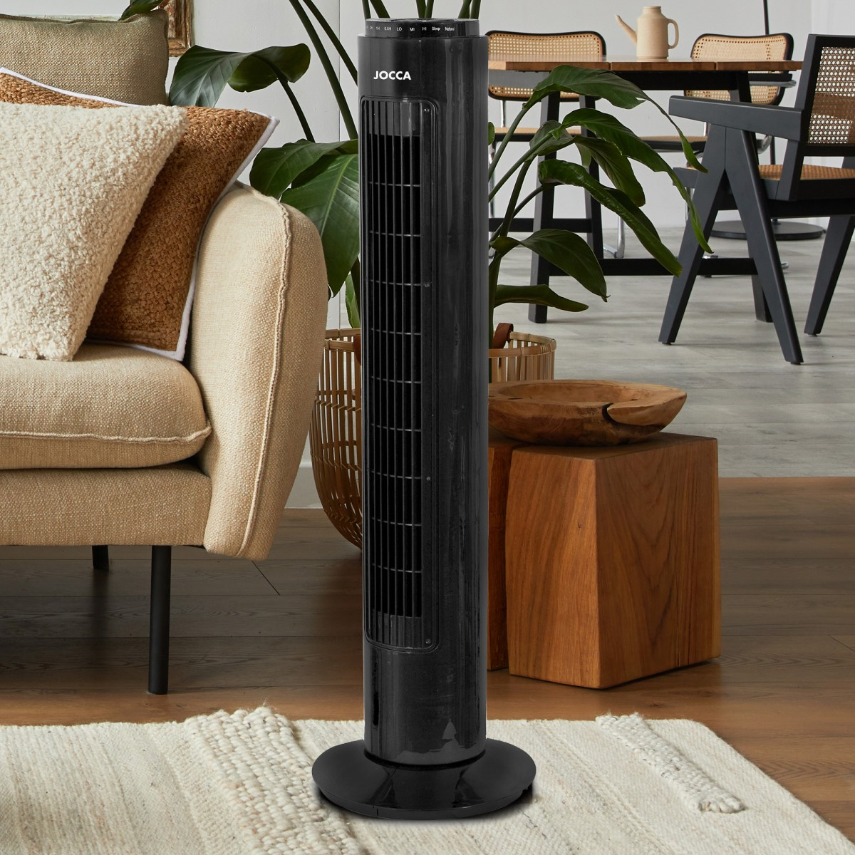Ventilador de torre negro en una habitación, con una mesa auxiliar de madera y un sofá beige.