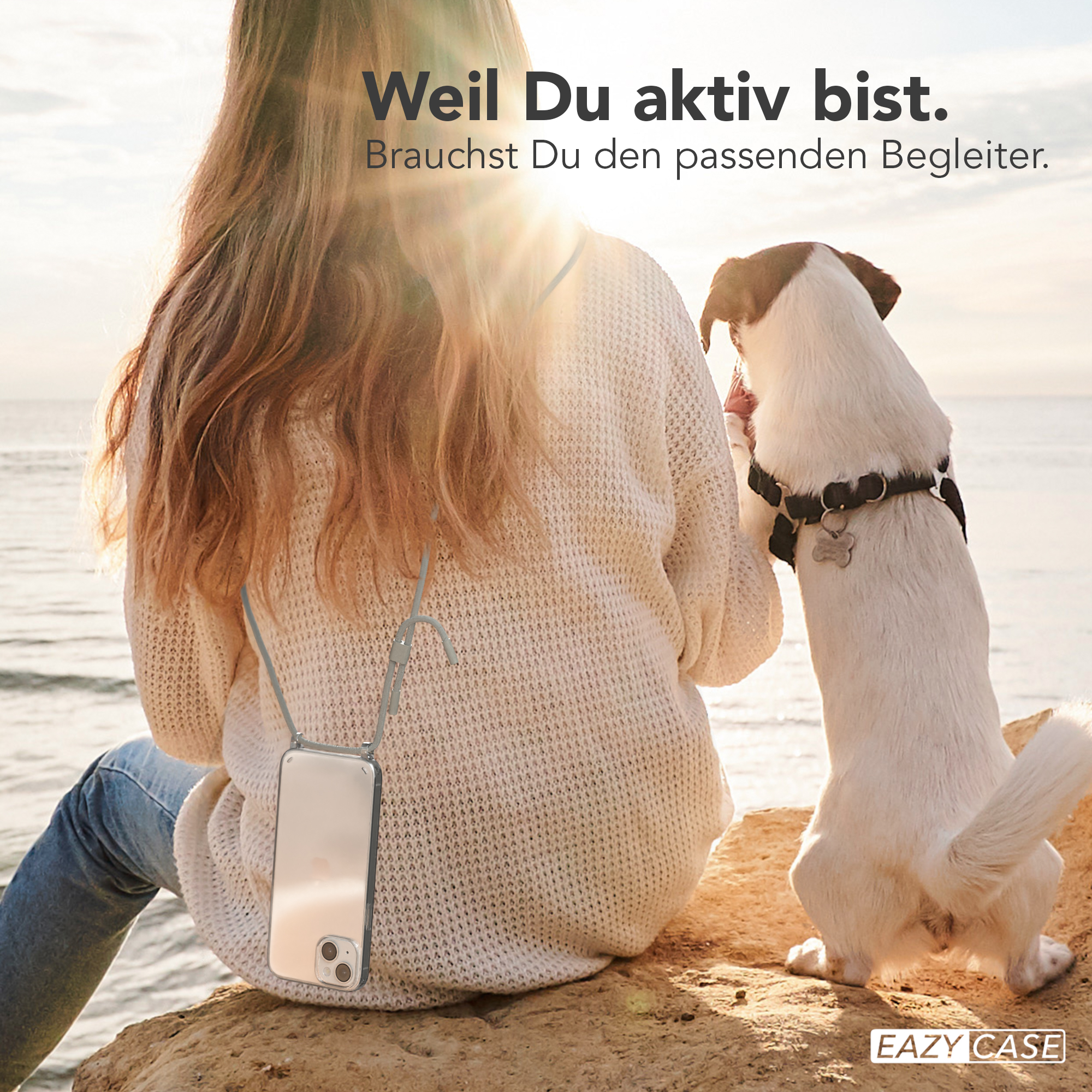 Eine Frau mit Telefon an einem Umhängeband sitzt auf einem Felsen und streichelt einen Hund. Sie sind am Meer.