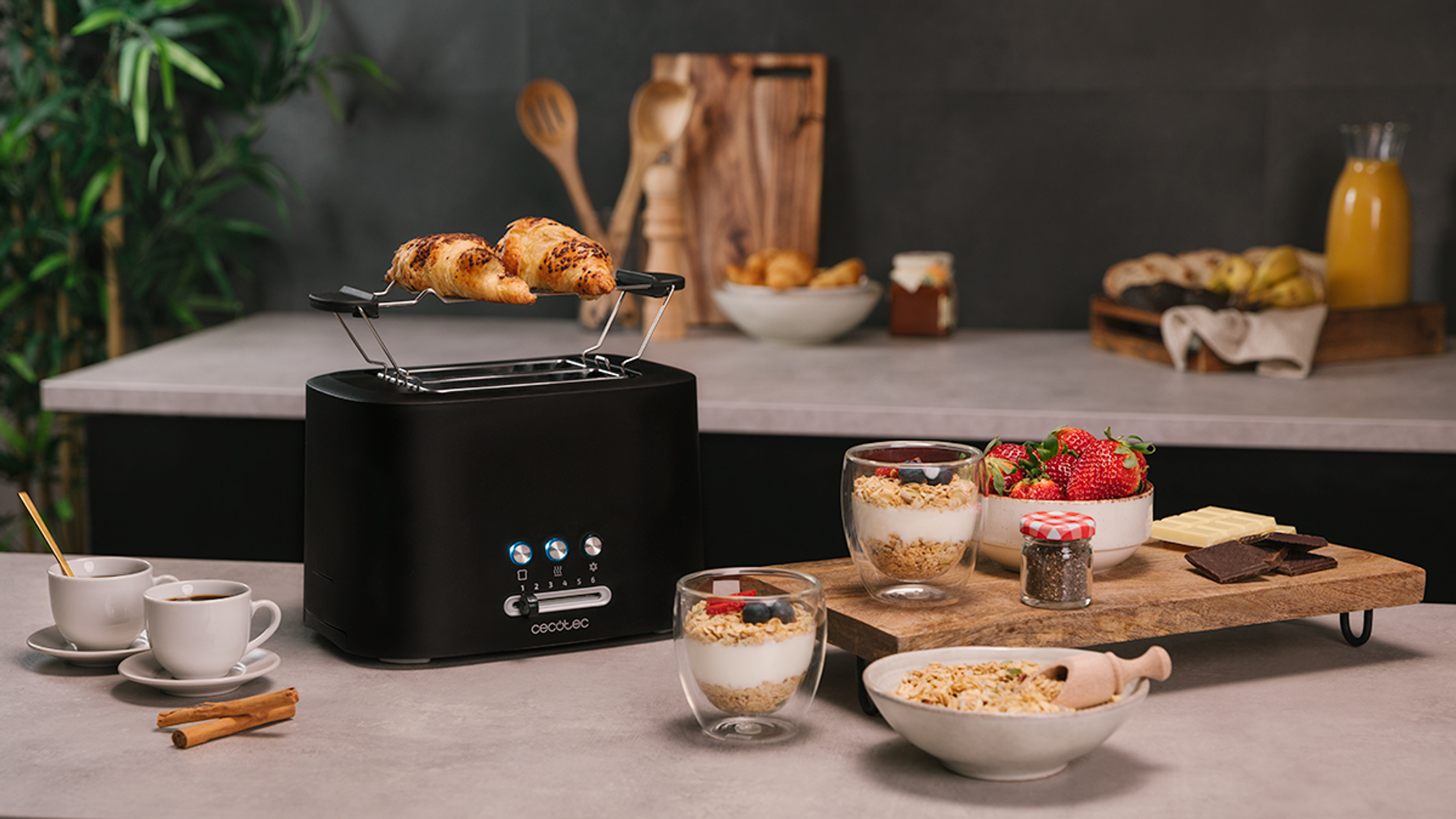 A black toaster with croissants on top and other breakfast items on the table. Includes yogurt and fruit.