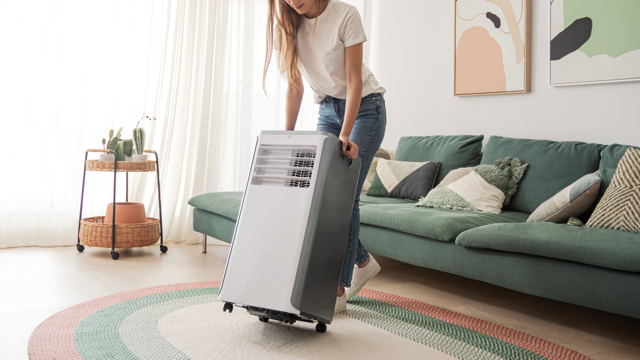 Kobieta toczy przenośny klimatyzator po dywanie na drewnianej podłodze. Zielona sofa w tle.