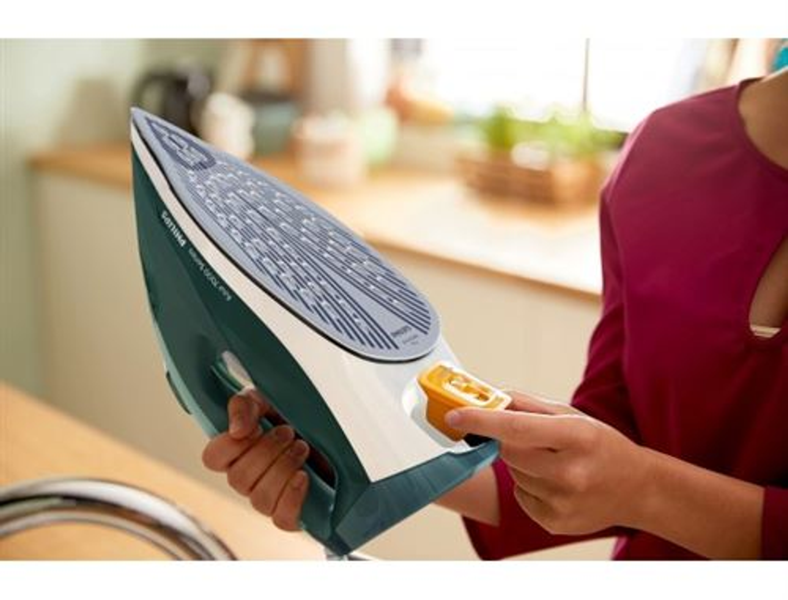 Osoba trzymająca żelazko. Zielono-białe żelazko z żółtym wlotem wody na drewnianej powierzchni.