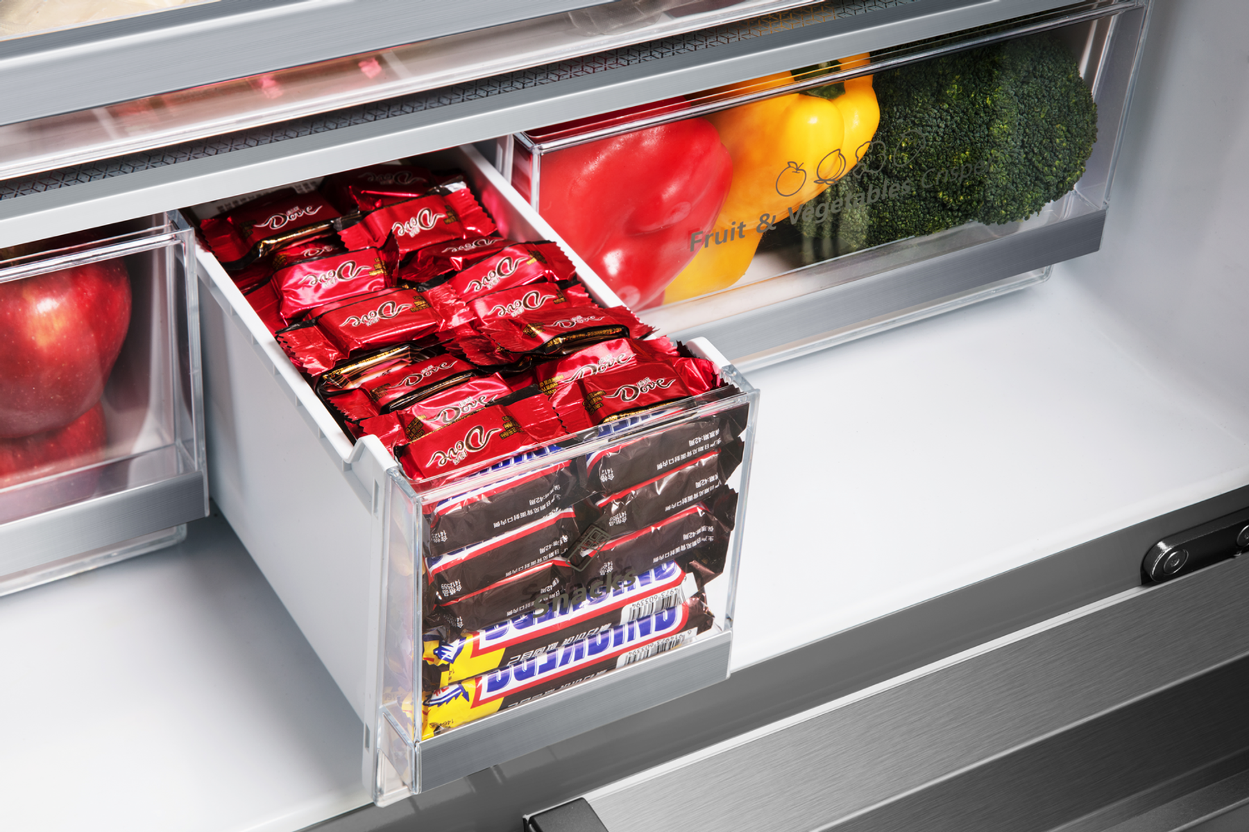 Cajón de refrigerador abierto lleno de barras de chocolate y verduras frescas.
