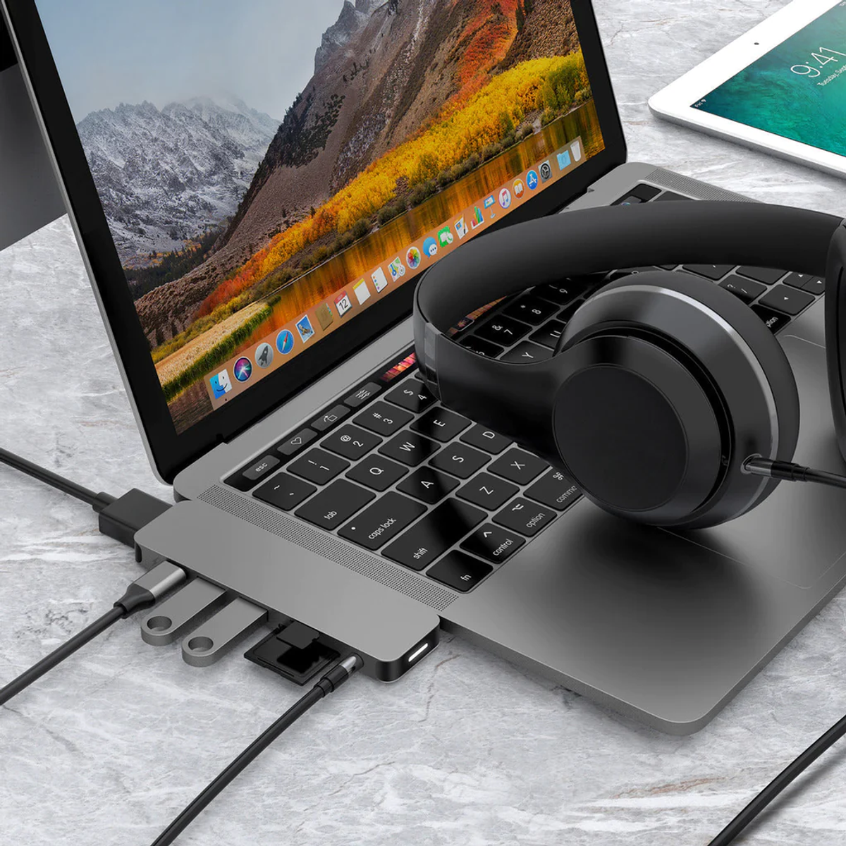 Un laptop con un hub collegato e cuffie. Un tablet è visibile sullo sfondo, vista montagna.