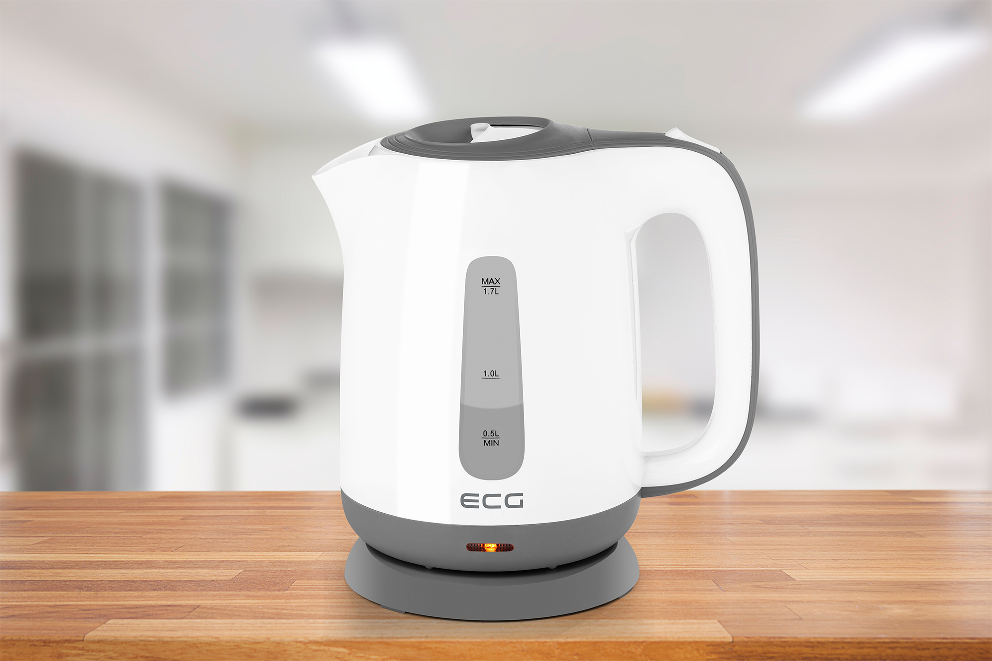 Obraz przedstawia biały czajnik elektryczny z szarą podstawą, na drewnianym stole, z rozmytym tłem kuchni.