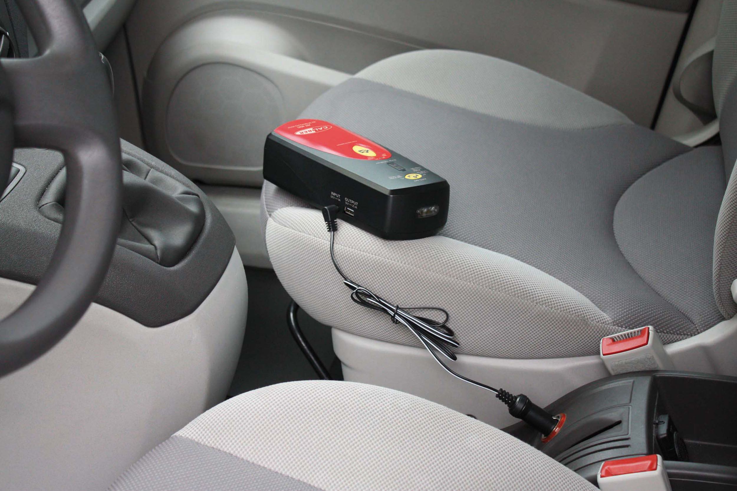Ein schwarz-roter Starthilfe-Booster auf einem Autositz, mit angeschlossenen Kabeln.