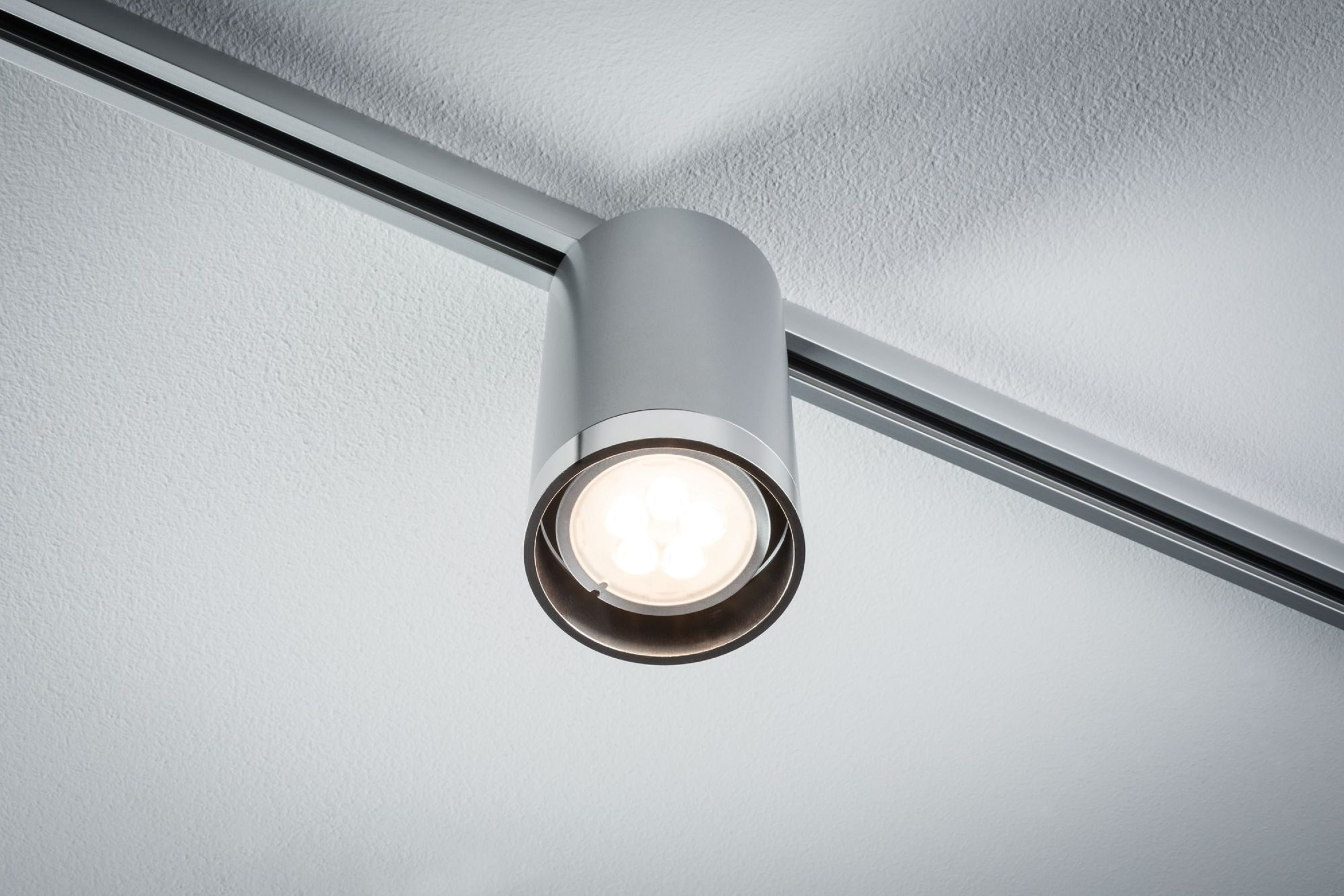 Silberner zylindrischer Strahler mit hellem Licht, montiert an einer Schiene an einer weißen Decke.