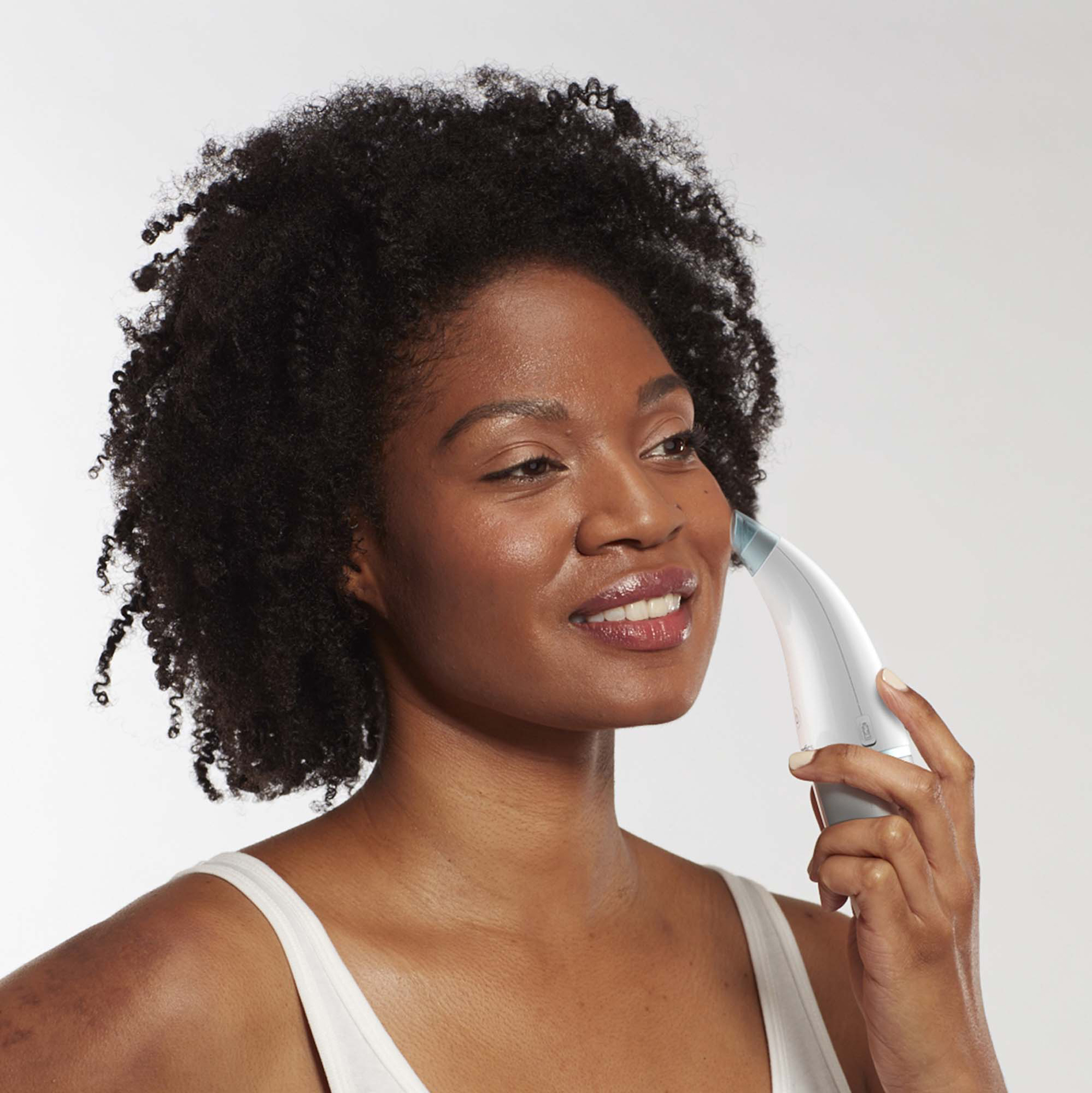 Kobieta używająca urządzenia do oczyszczania twarzy. Ma ciemne kręcone włosy i uśmiecha się.