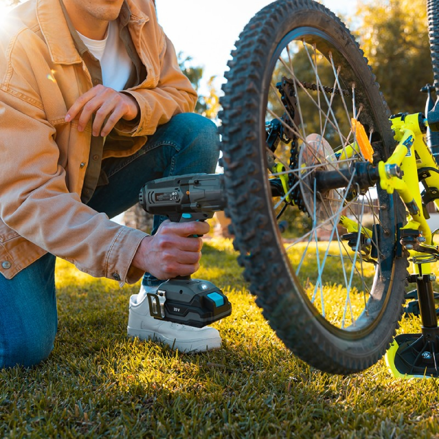 Mężczyzna klęczy na trawie, używając wiertarki do koła roweru, aby coś dokręcić.
