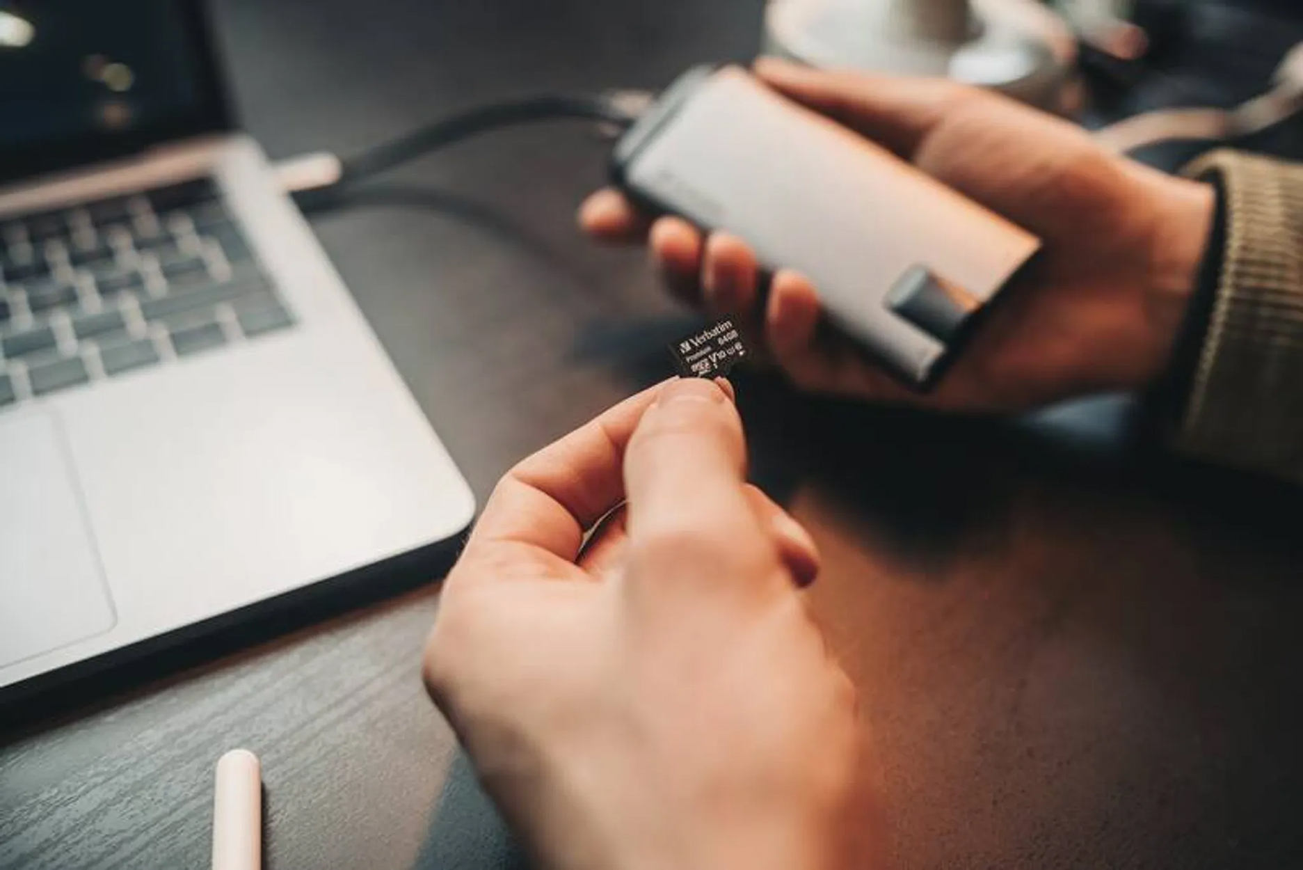 Dłonie osoby trzymającej kartę SD i urządzenie, w pobliżu laptopa na drewnianym biurku.