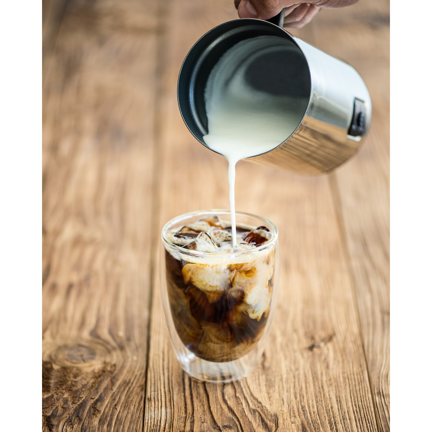 Milch wird in ein Glas Eiskaffee auf einem Holztisch gegossen.