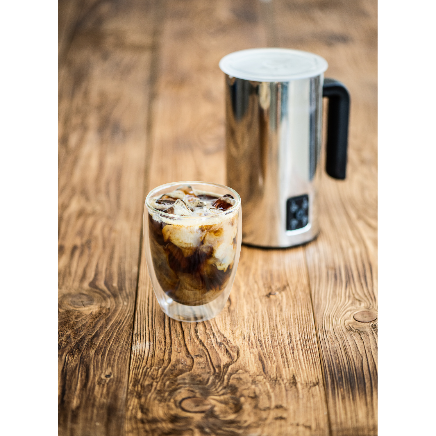 Glas Eiskaffee neben silbernem Milchaufschäumer auf Holz.