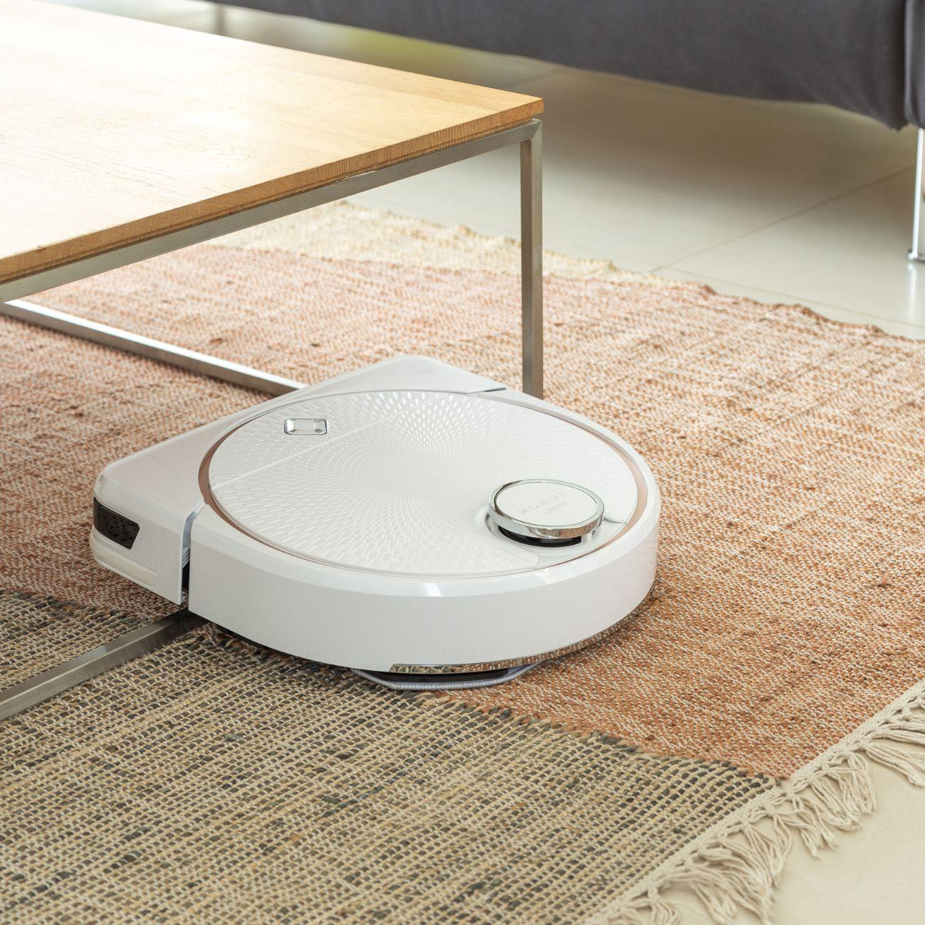 Aspiradora robot blanca sobre una alfombra debajo de una mesa. La alfombra es marrón y beige.