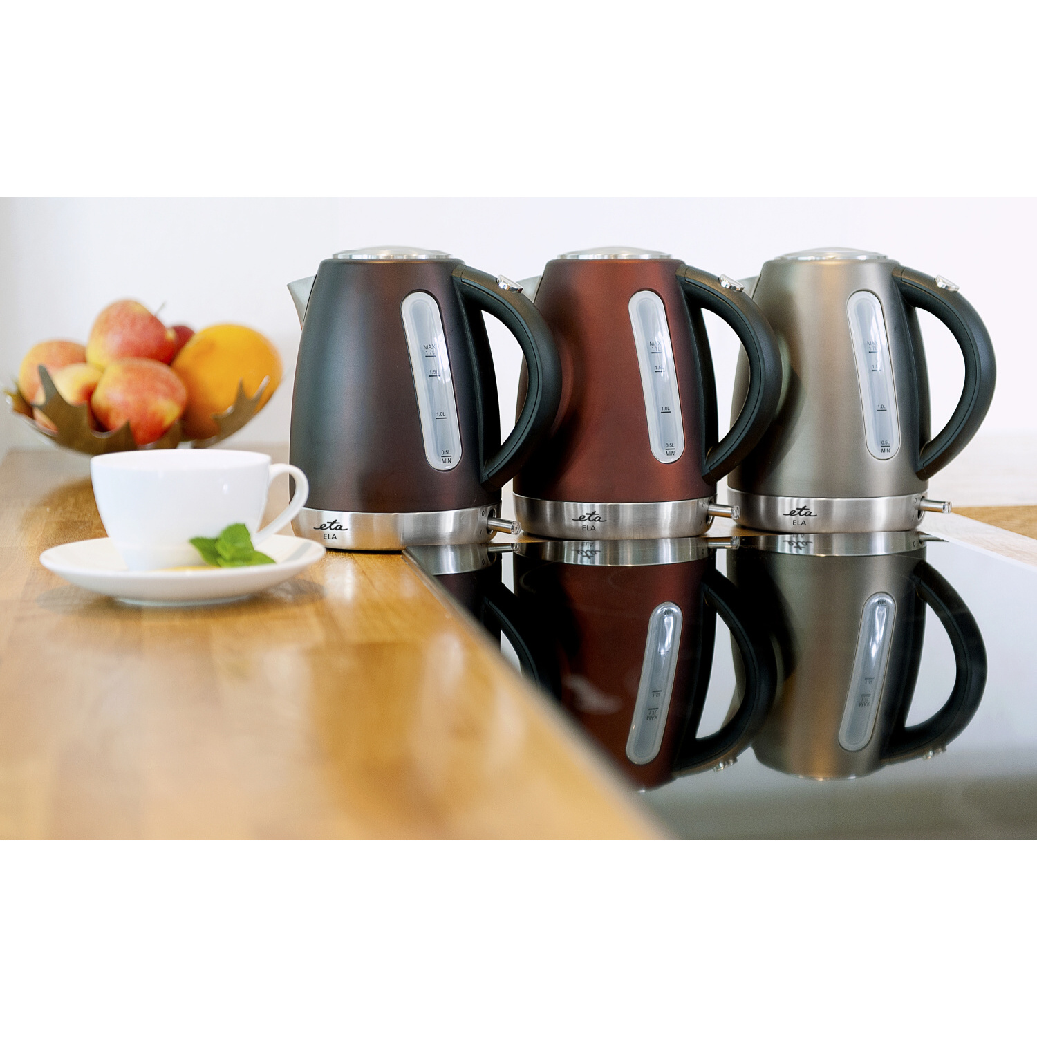 Drei Wasserkocher in Braun, Rot und Silber, mit einer weißen Tasse und einer Obstschale.
