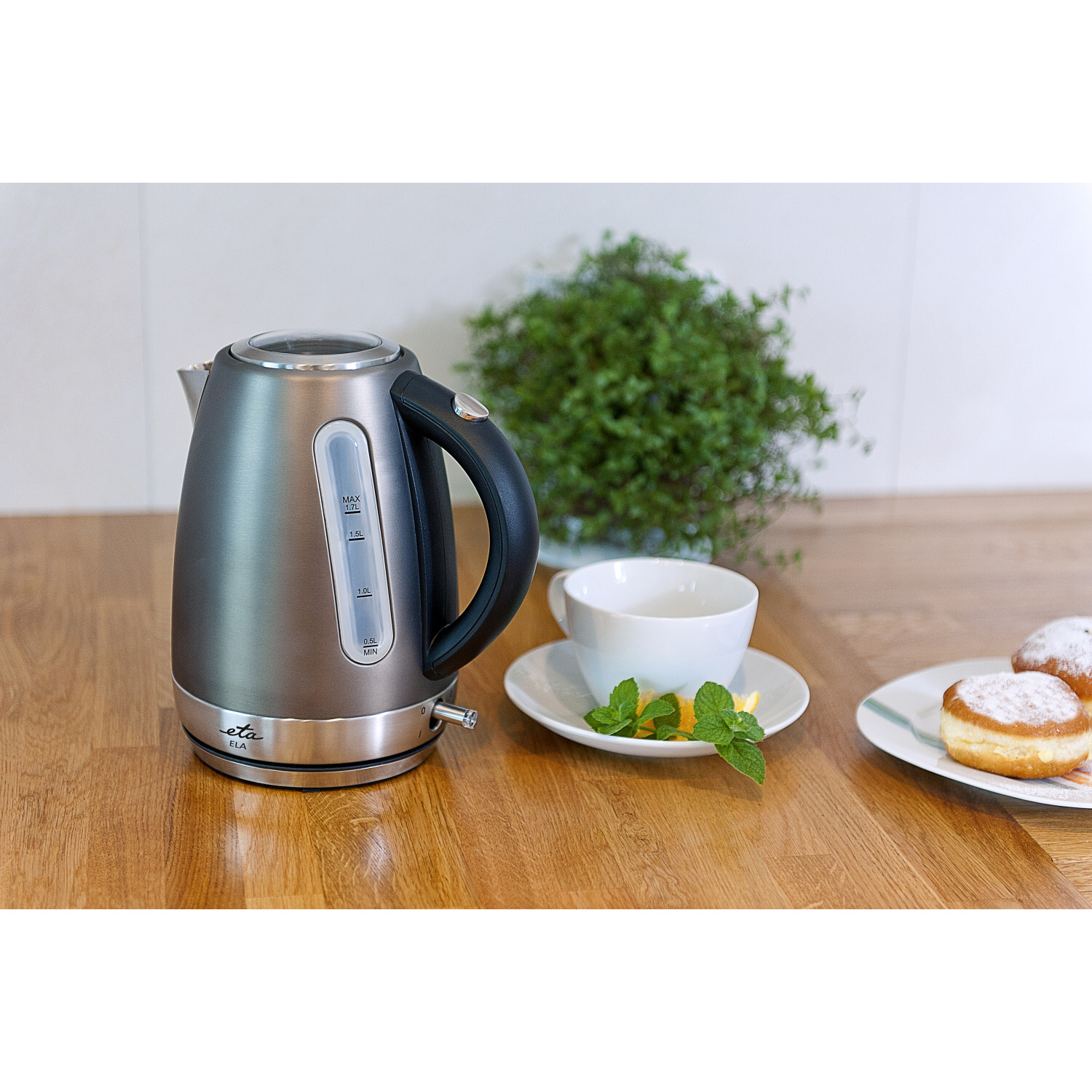 Hervidor eléctrico de acero inoxidable con taza, rosquillas y planta en mostrador.