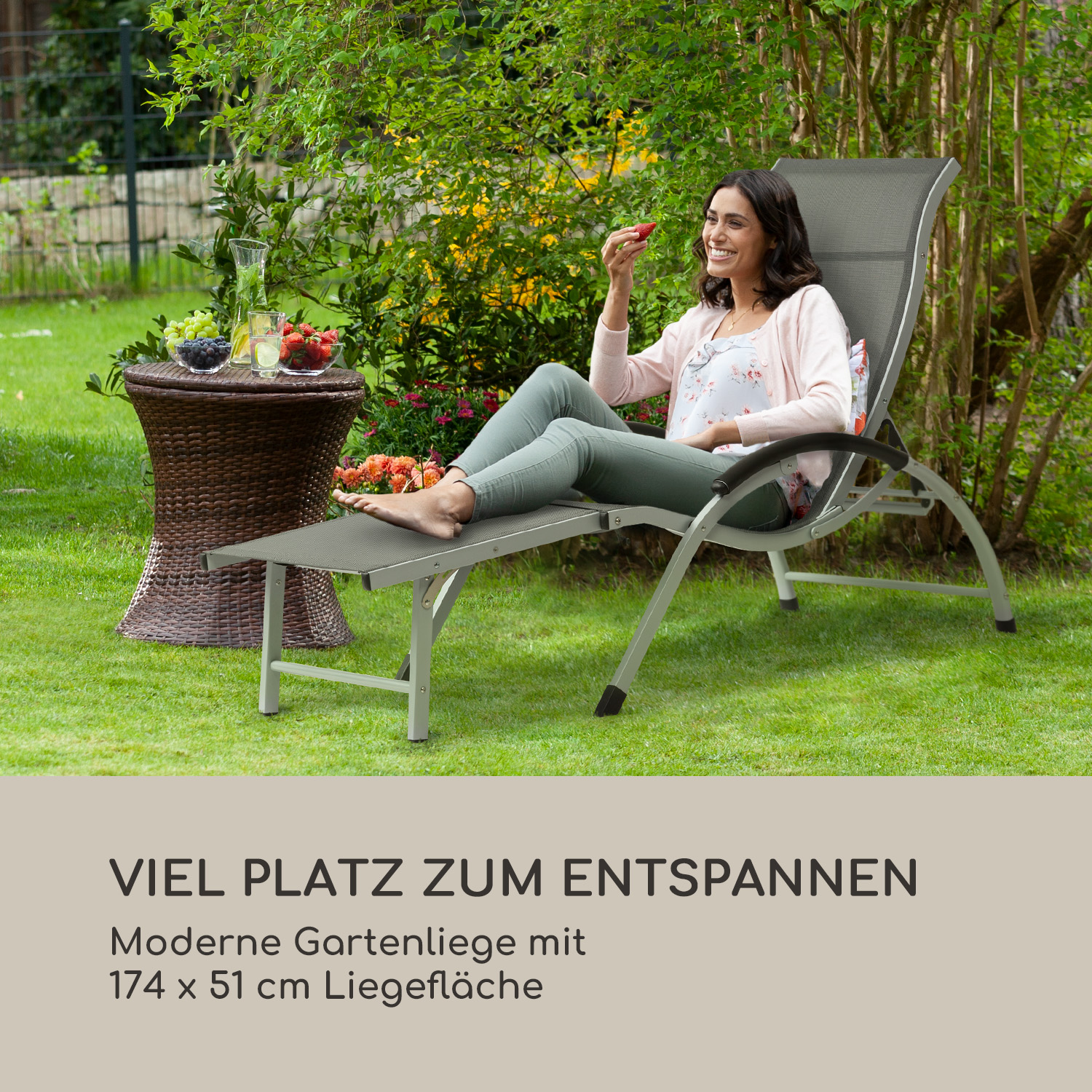 Eine Frau sitzt auf einem grauen Liegestuhl im Garten und hält eine Erdbeere.
