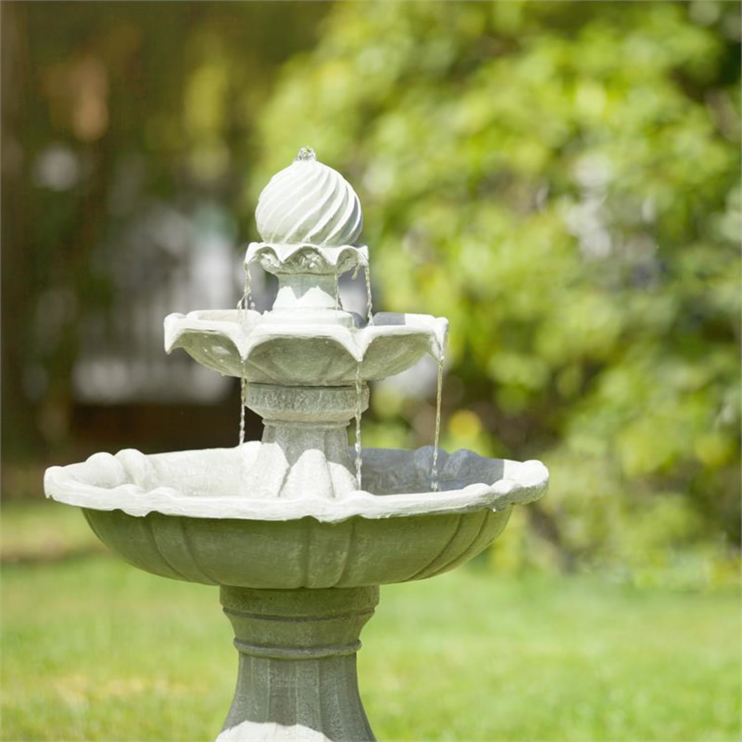 Ein Steinfontein in einem Garten. Wasser fließt von der oberen zur unteren Ebene. Grünes Laub im Hintergrund.
