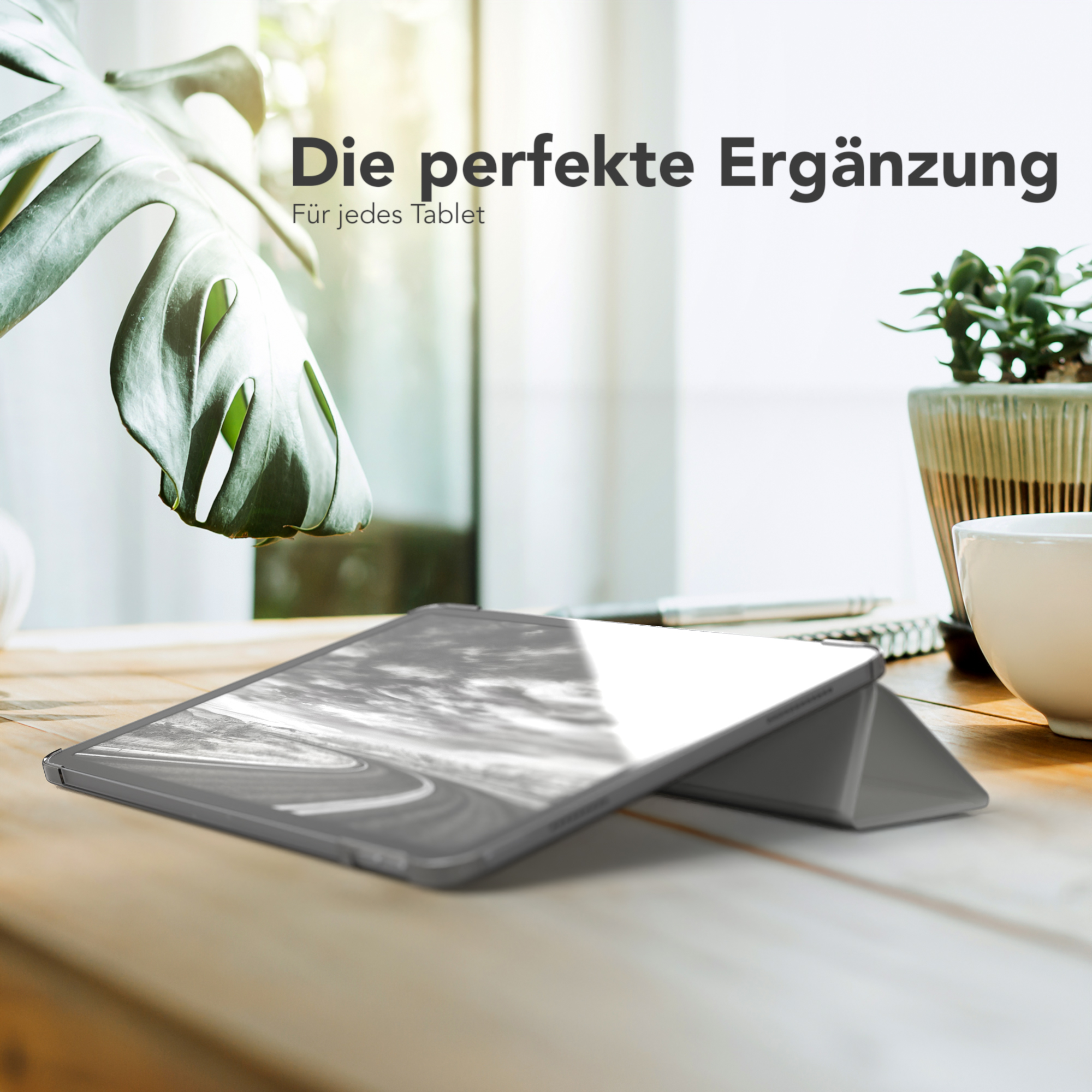 Tablet in einem grauen Gehäuse auf einem Holztisch, mit Fenster und Pflanze im Hintergrund.