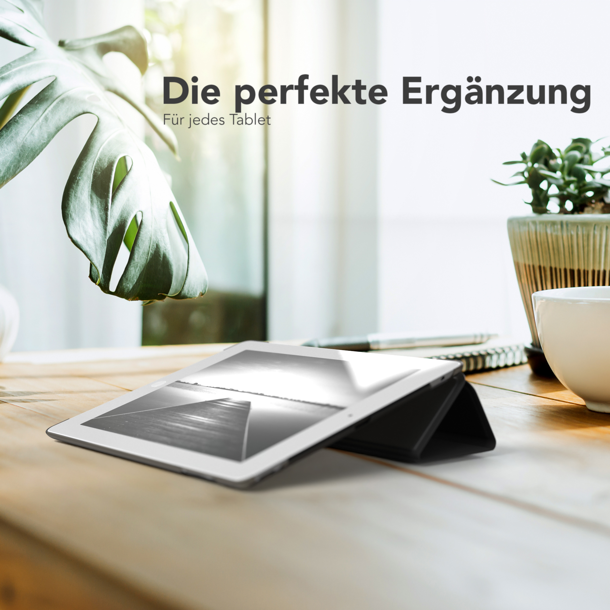 Ein Tablet liegt auf einem Holztisch. Eine grüne Pflanze und eine weiße Tasse stehen daneben. Das Tablet hat schwarze Abdeckung.