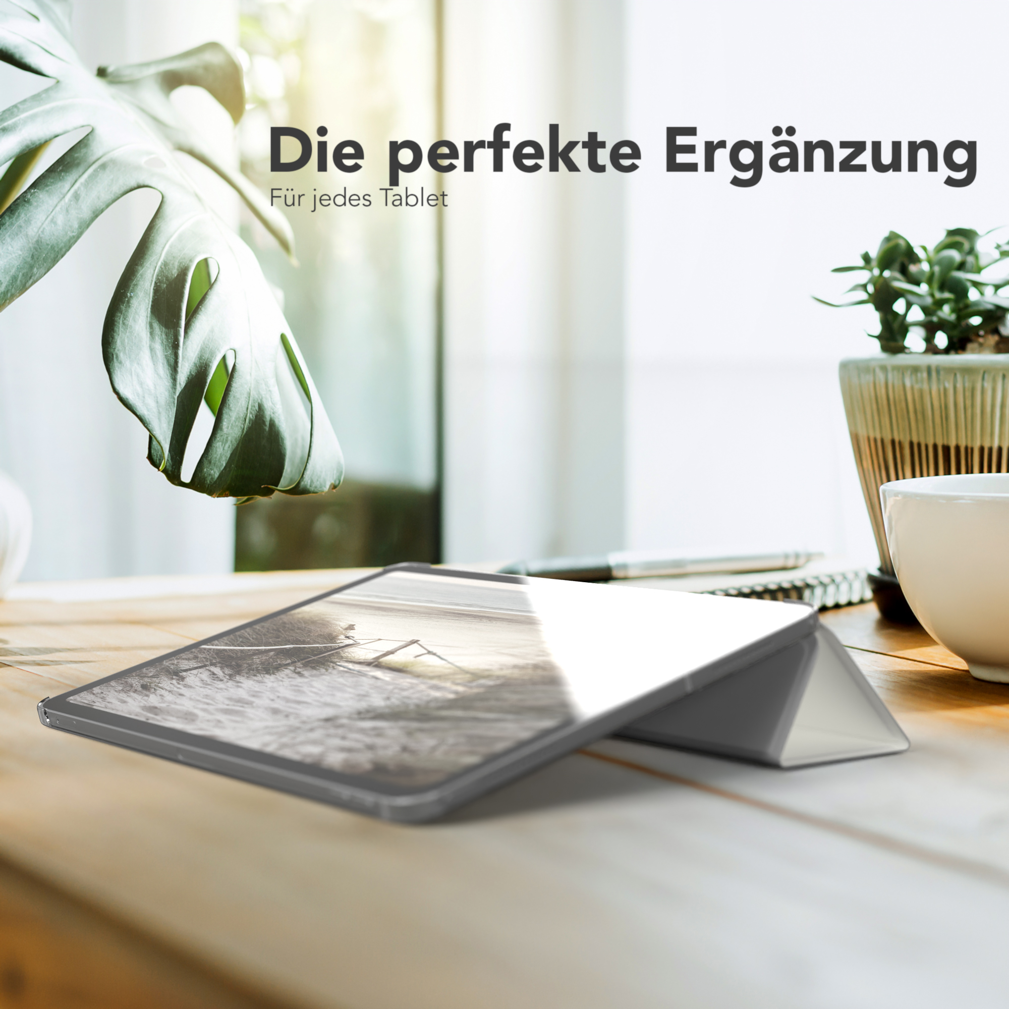 Ein Tablet mit Hülle auf einem Holztisch. Eine Pflanze und eine Tasse sind im Hintergrund.